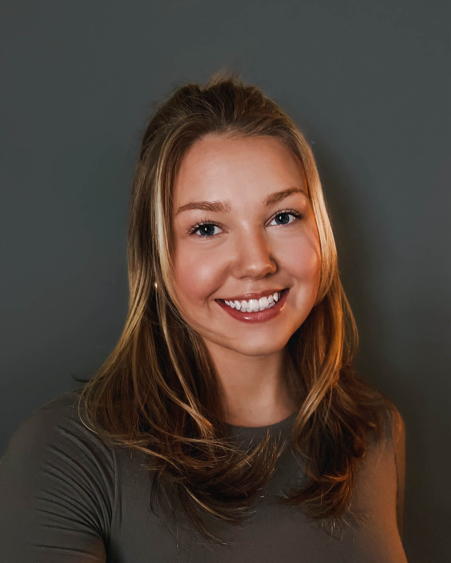 Portrait photo against dark gray background showing a bright smile and long brown hair.