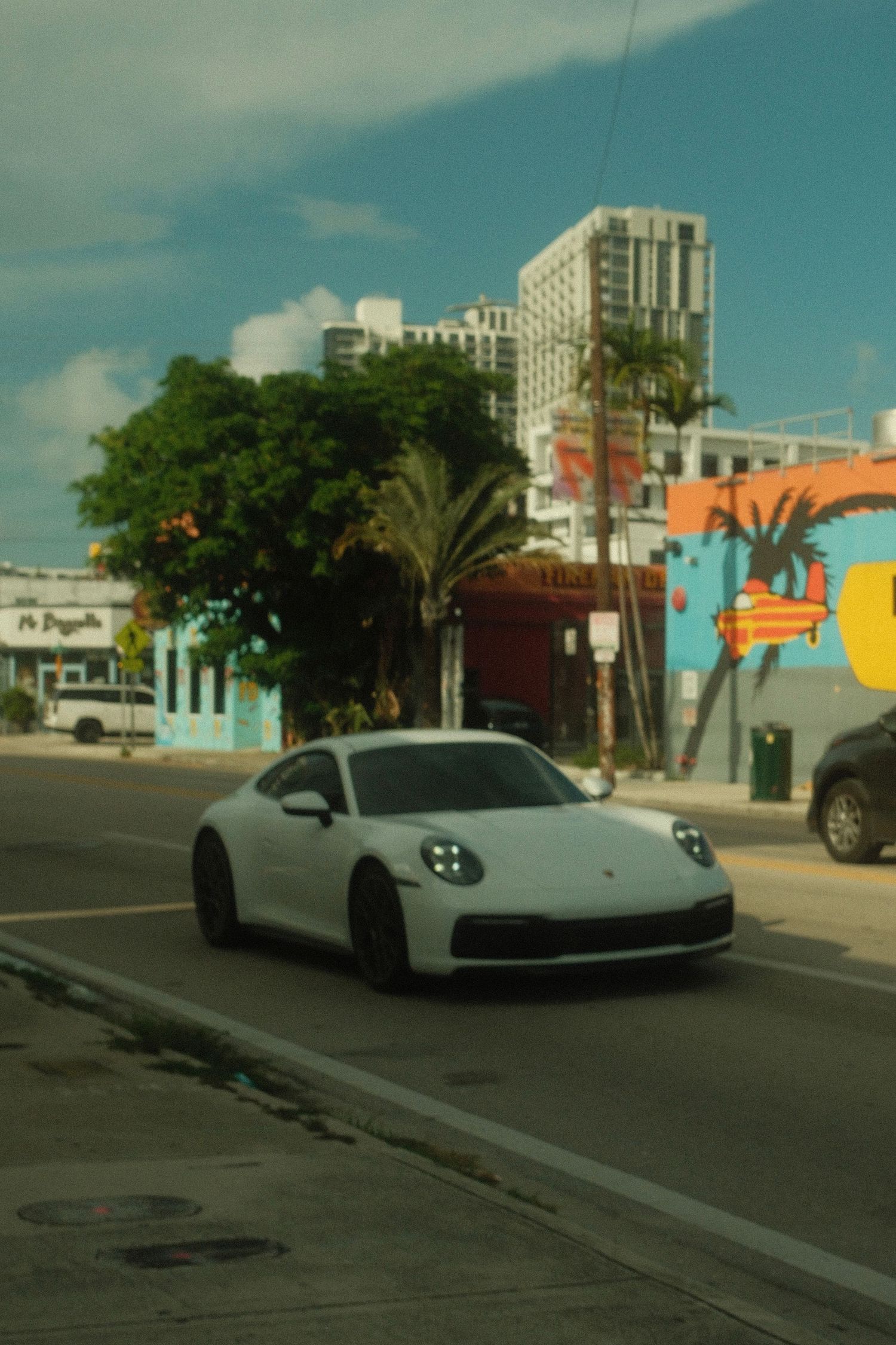 White Porsche 911 sports car parked on a Miami street with palm trees and buildings in the background.