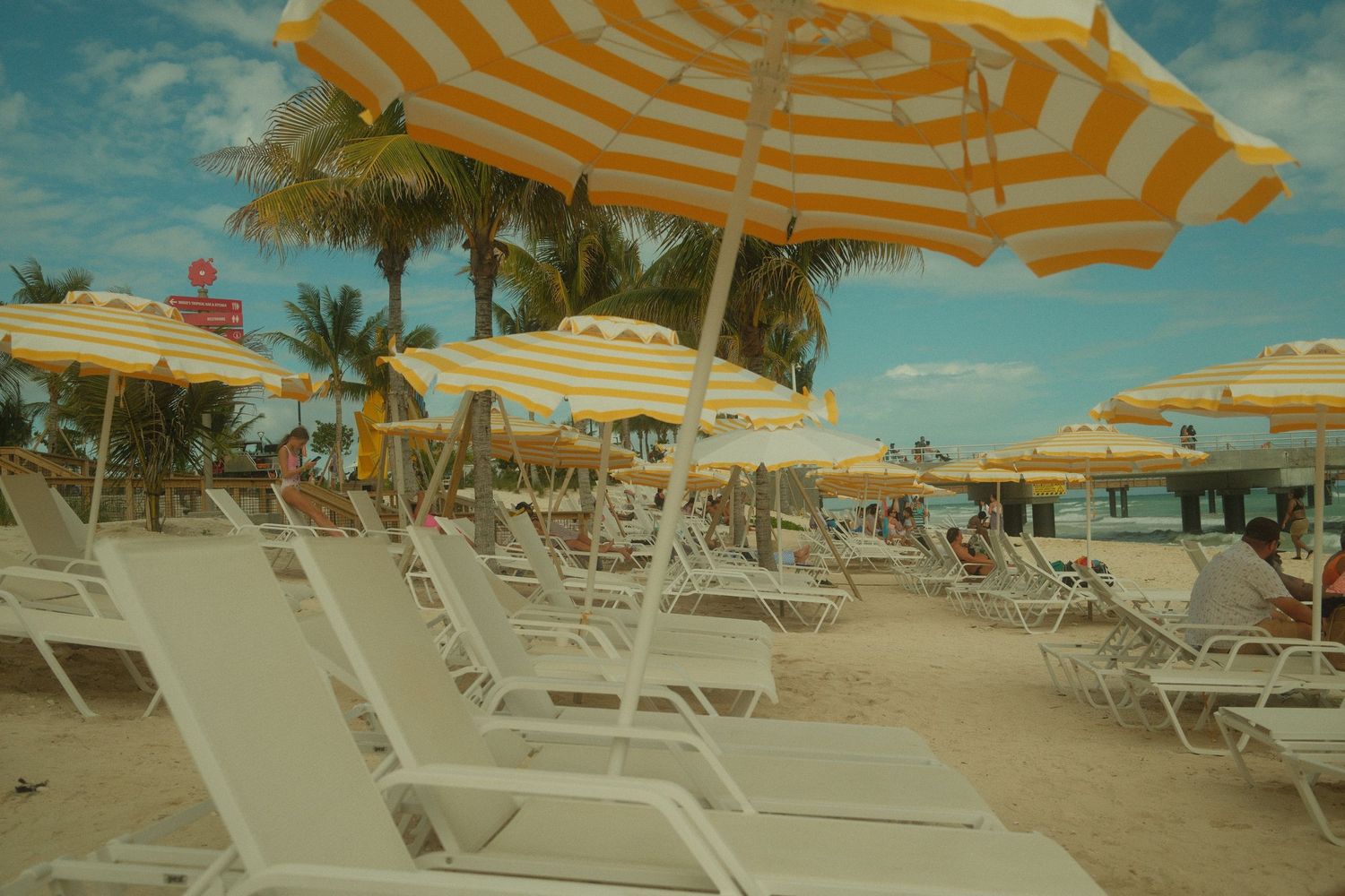 Orange and white striped beach umbrellas shade lounge chairs on a sunny tropical beach with palm trees.