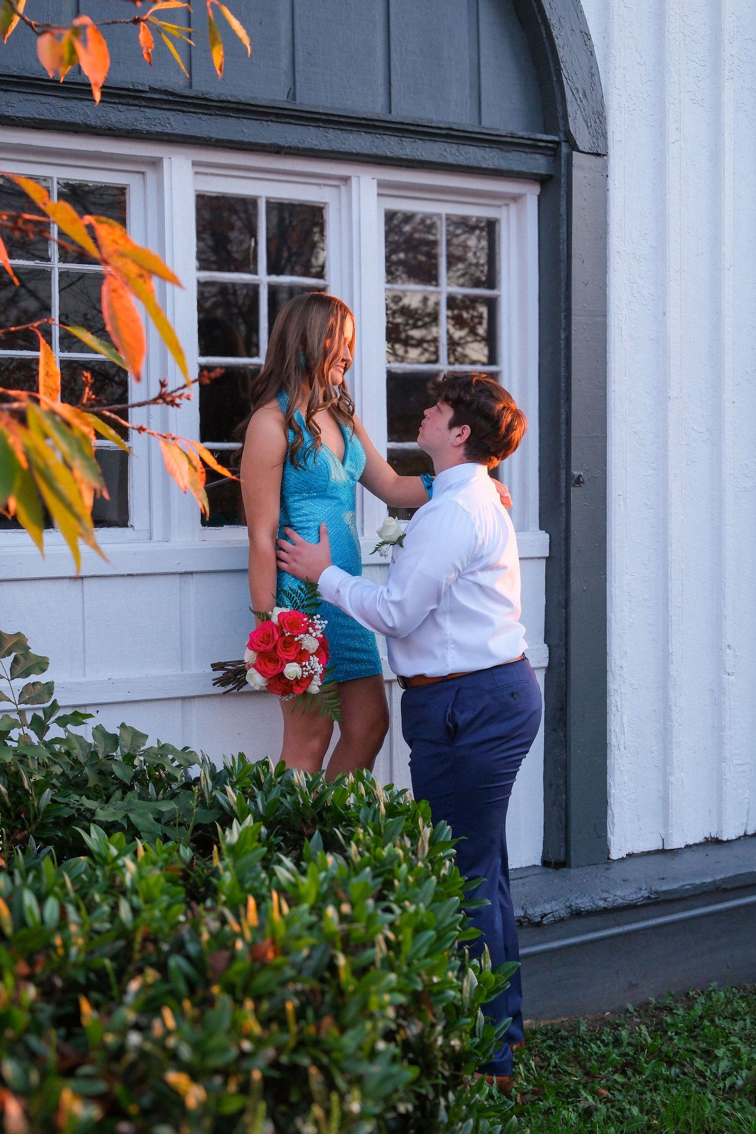 A couple shares a romantic moment outside a white building with green trim at sunset.