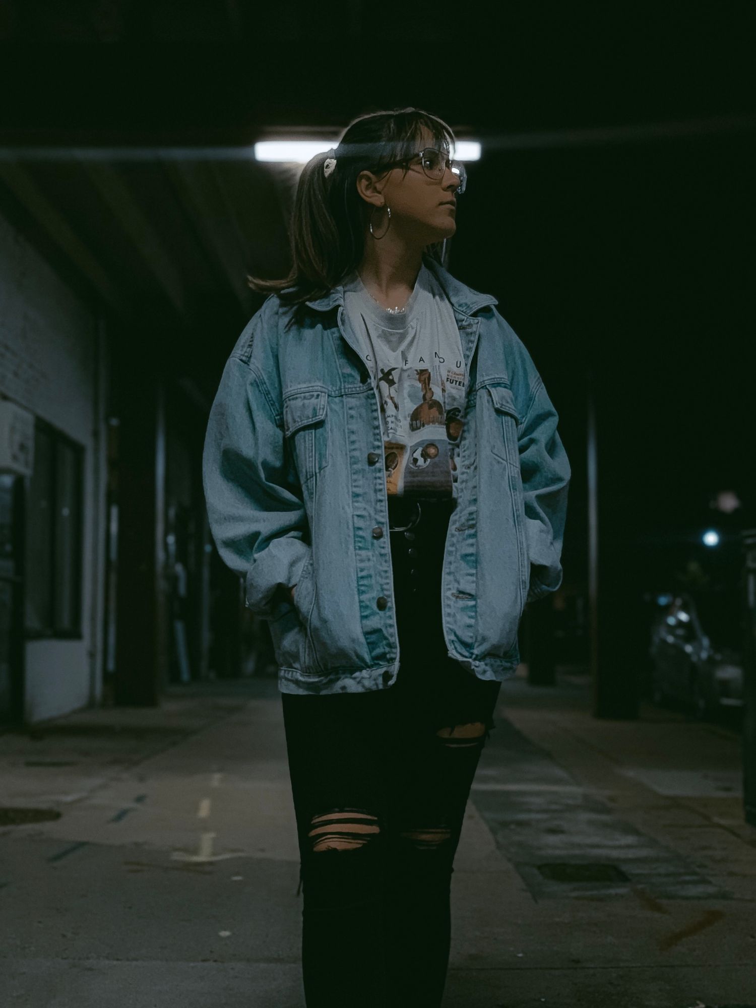 Person in oversized denim jacket and ripped black jeans poses on dark street at night.