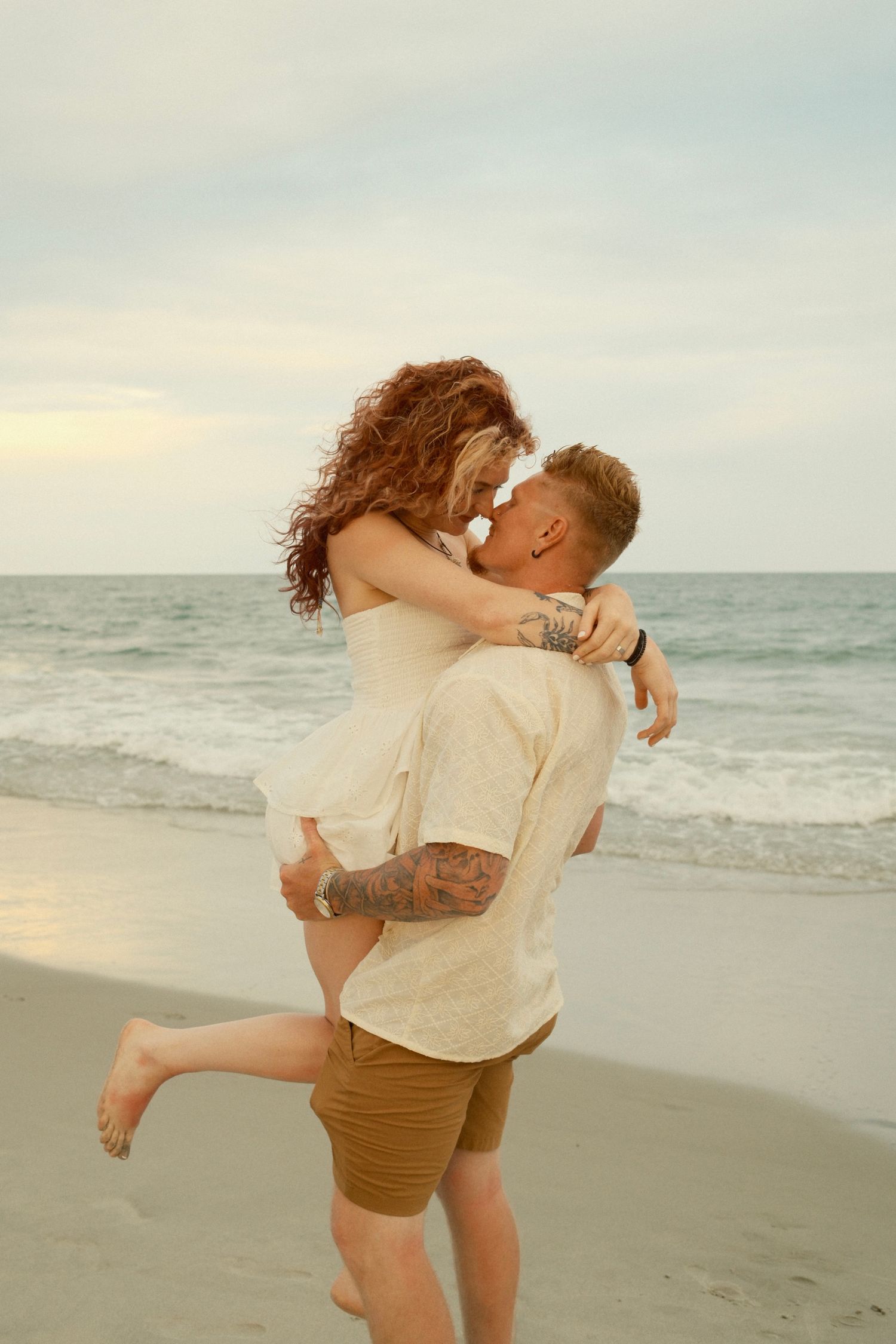 A couple shares an intimate moment on the beach shore with soft lighting and crashing waves behind them.