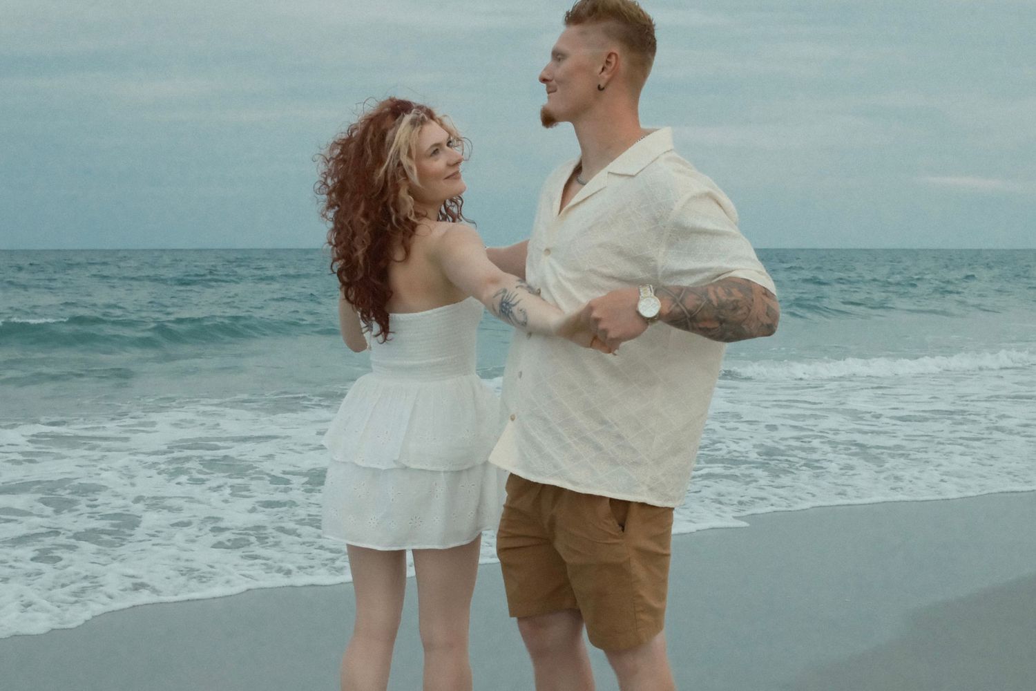 Couple in light colored clothing dancing together on beach with waves rolling in.