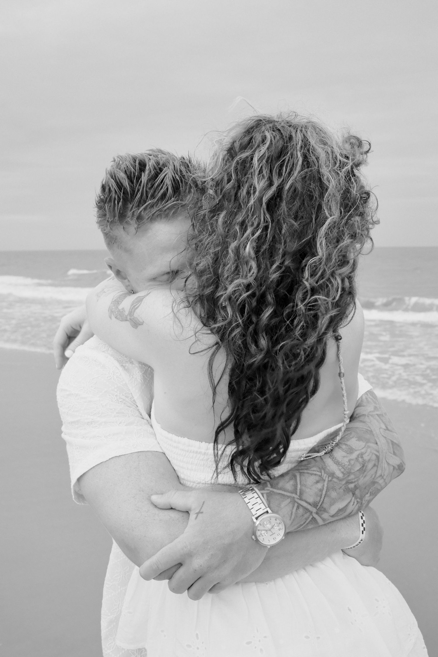 A romantic couple in white embraces and kisses on a beach with ocean waves in the background.