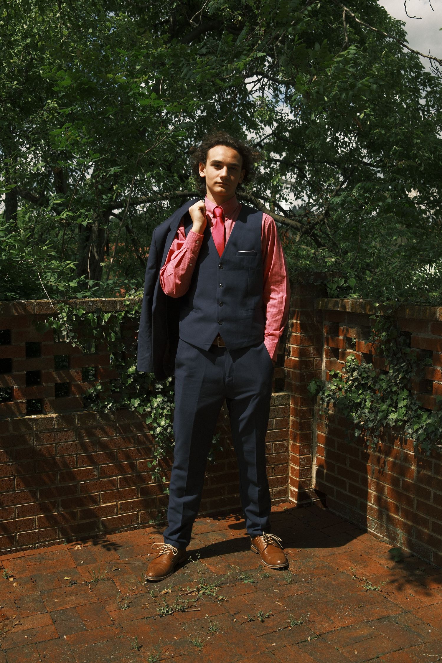 Person in navy vest and red shirt poses against brick wall in outdoor garden setting.