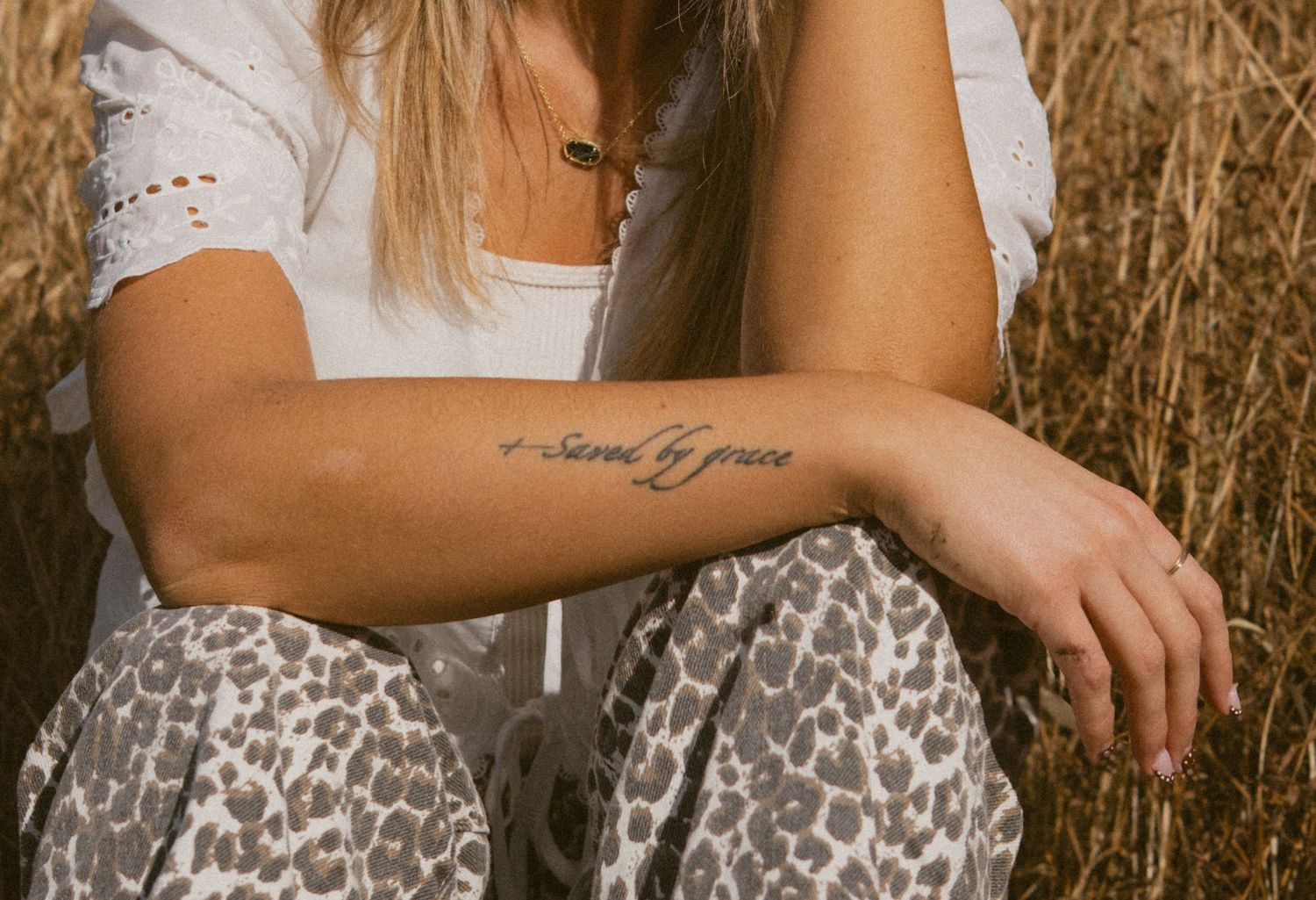Woman in white top and leopard print skirt stands in tall golden grass during sunset.