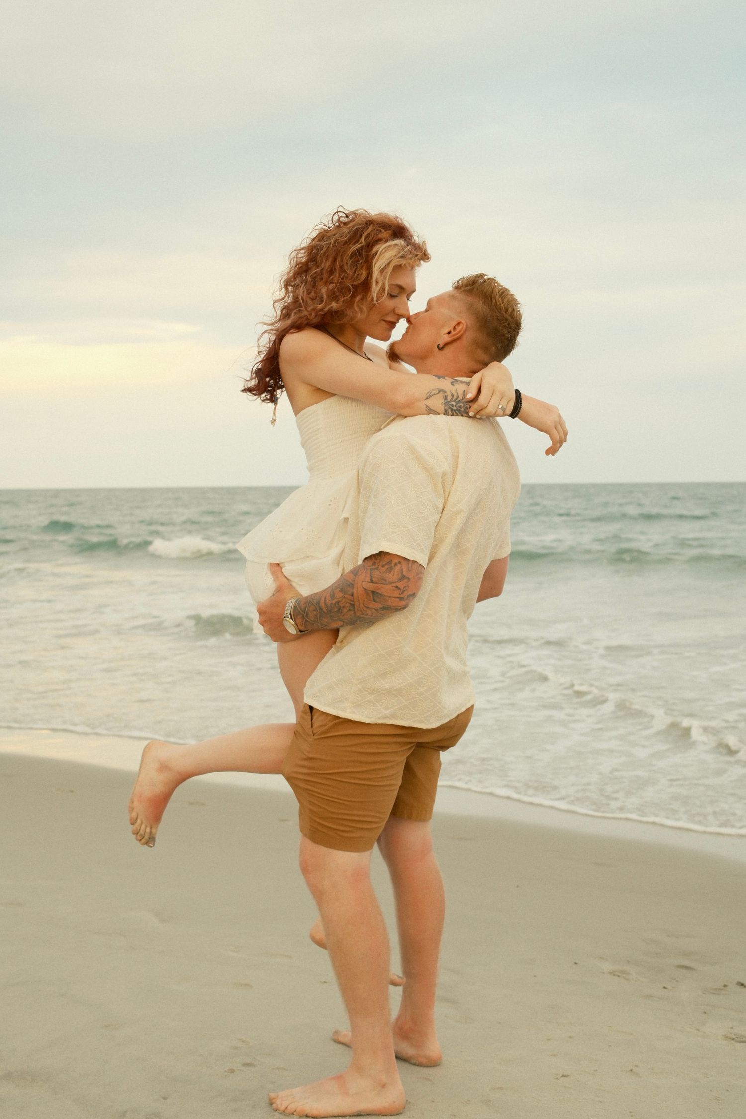 A romantic couple embraces on the beach at sunset with ocean waves in the background.