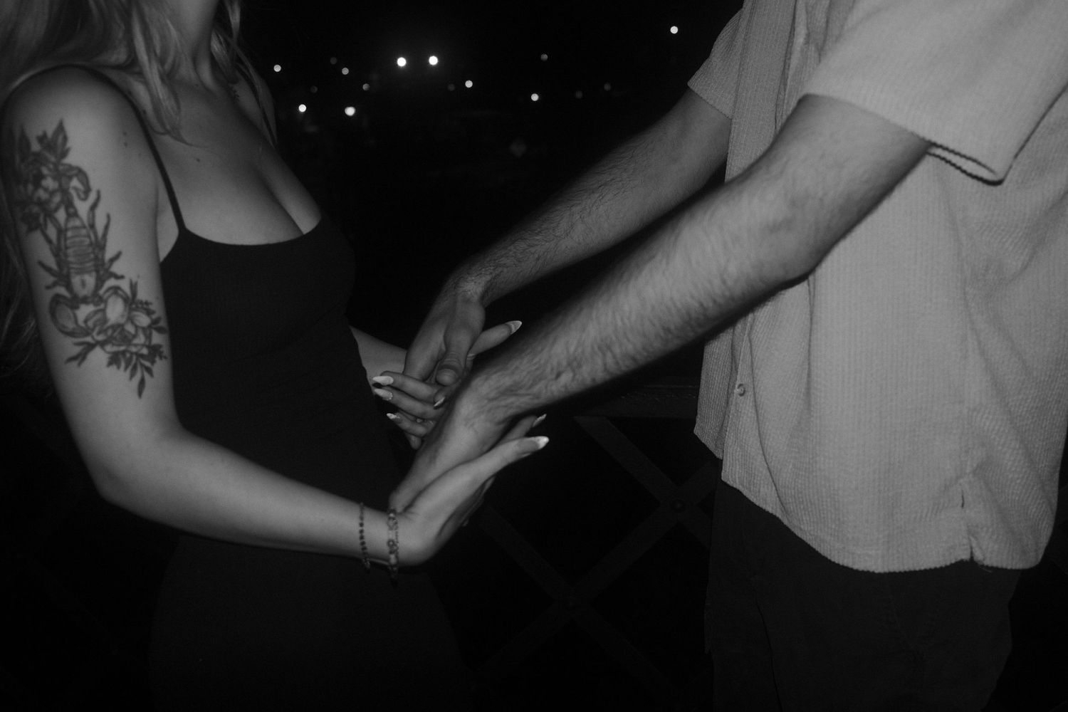 A black and white close-up photo shows two people holding hands with a visible arm tattoo in the dark.