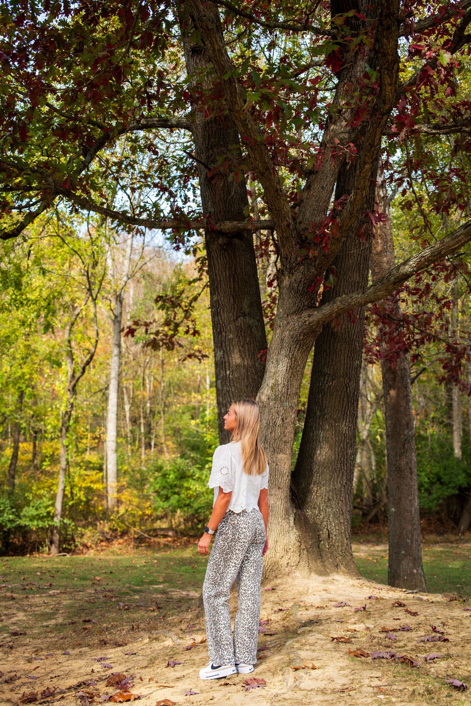 A figure stands against autumn trees in a wooded area wearing patterned pants and a white top.