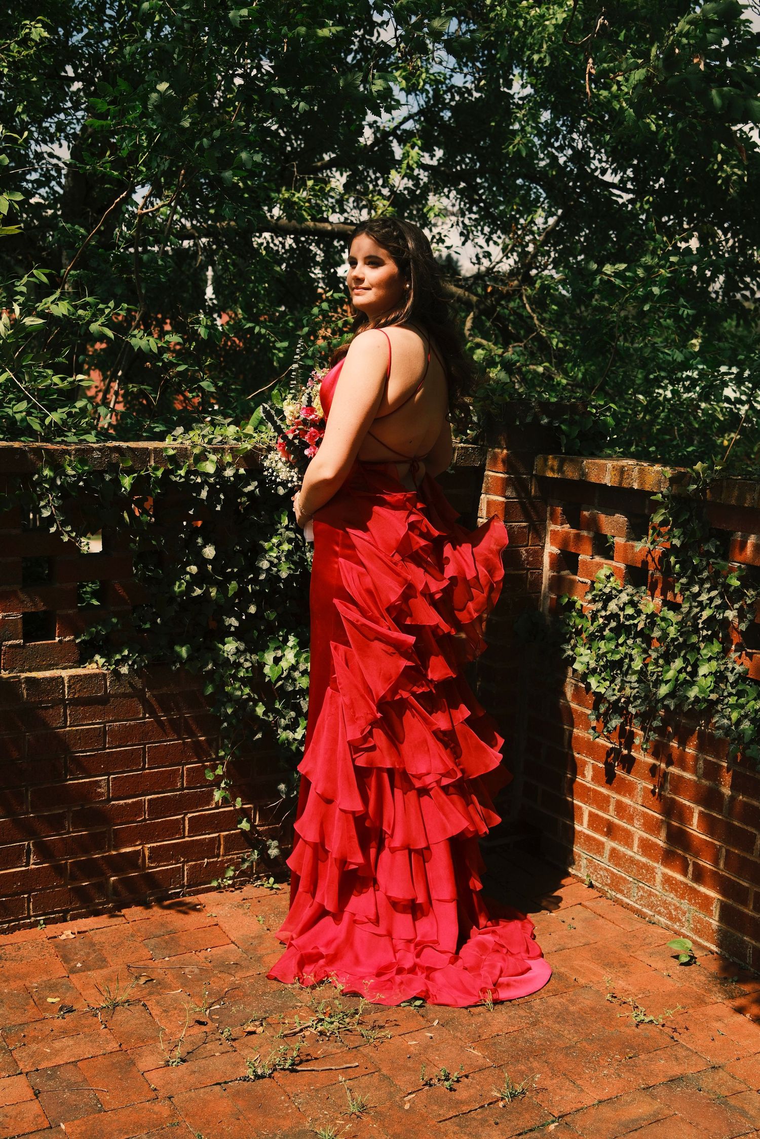 A person in a flowing red ruffled dress poses gracefully against a brick wall surrounded by lush greenery.