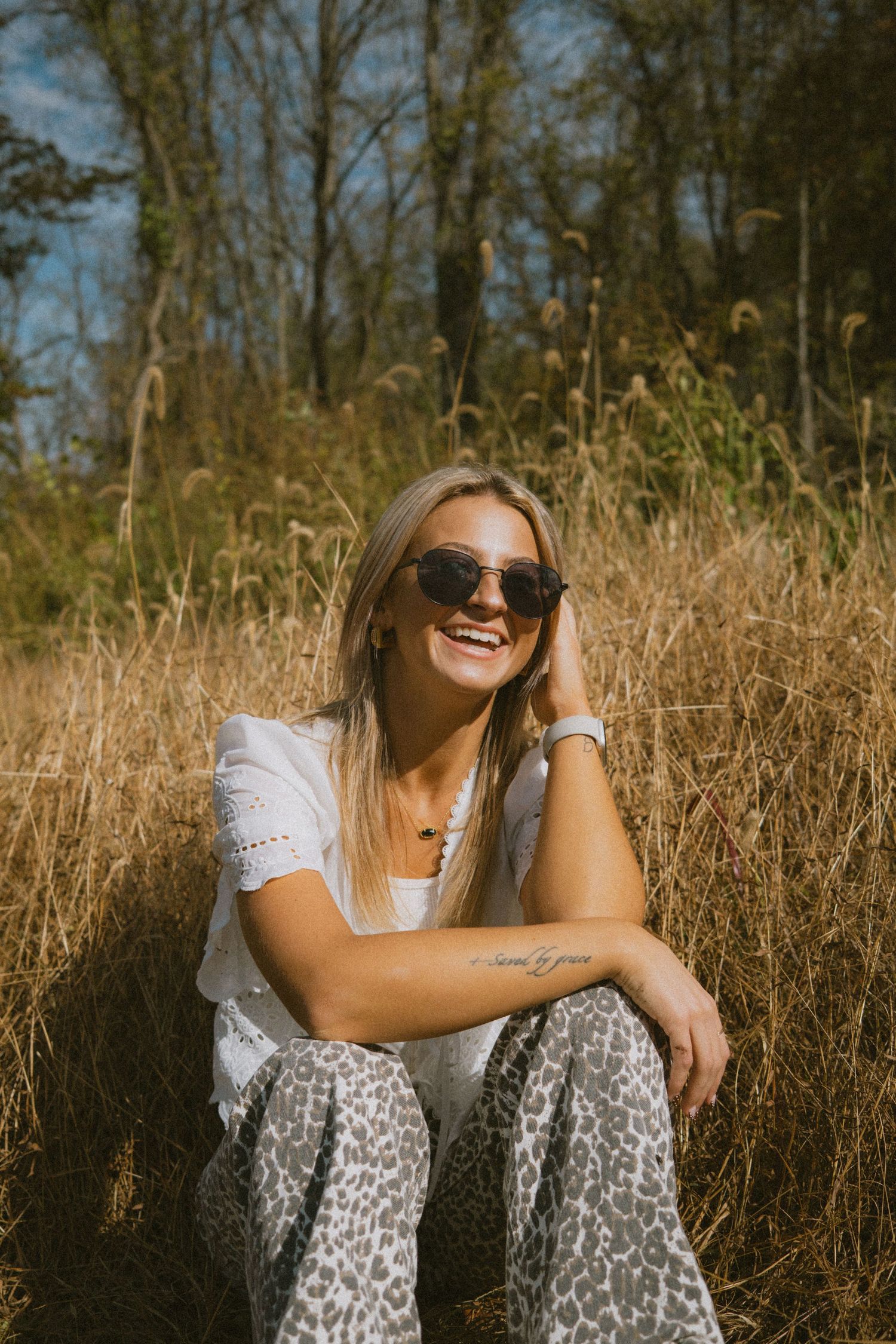 A woman in a white top and leopard print pants sits in golden grass during sunset, smiling with sunglasses.