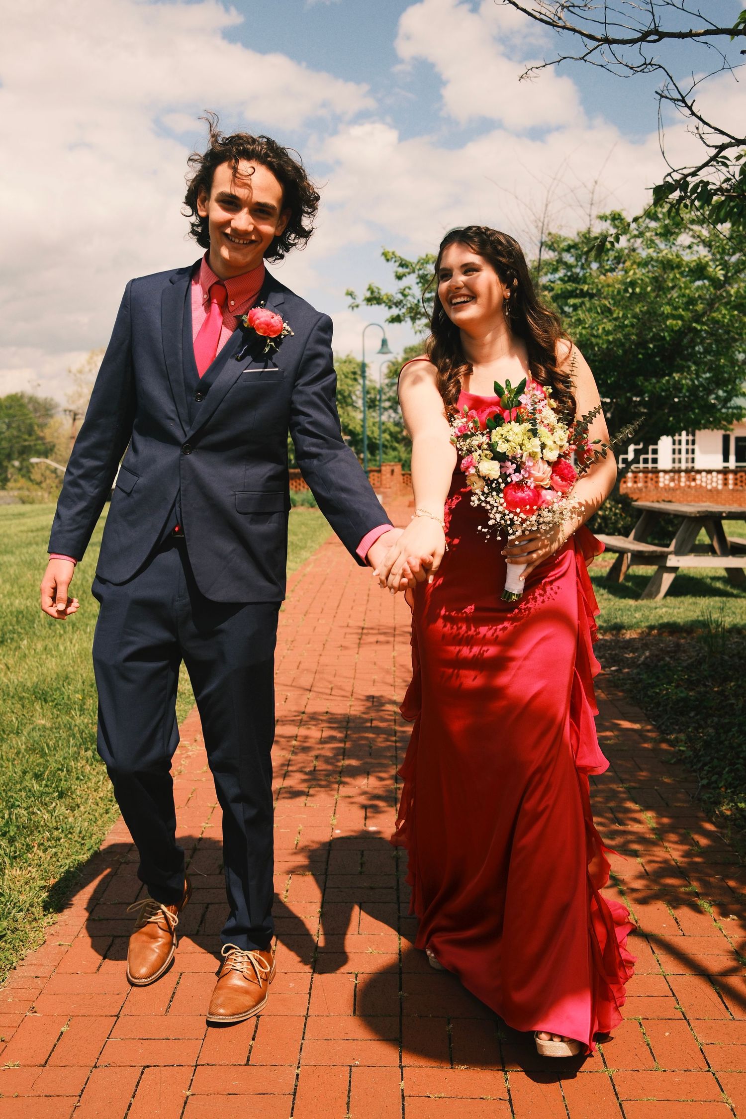 A couple in formal attire, with a navy suit and red dress, share playful moments on brick pavement outdoors.