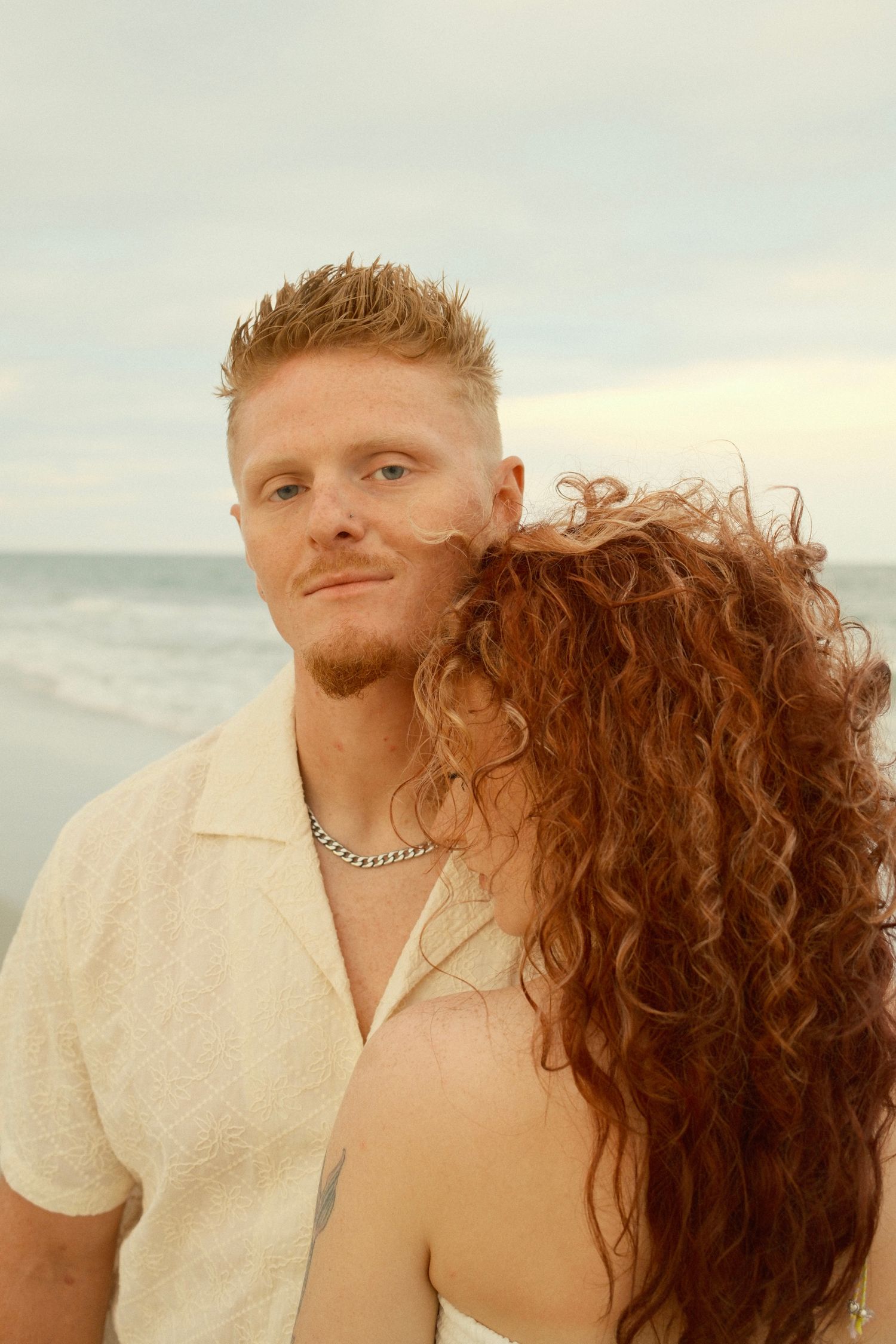 A romantic couple embraces on the beach during golden hour with the ocean behind them.