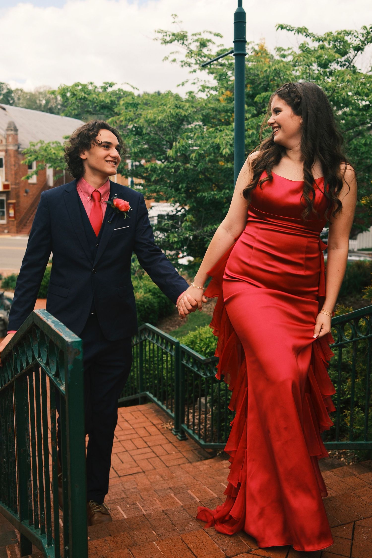 A couple in red and navy formal attire interact playfully on brick steps with green railings.