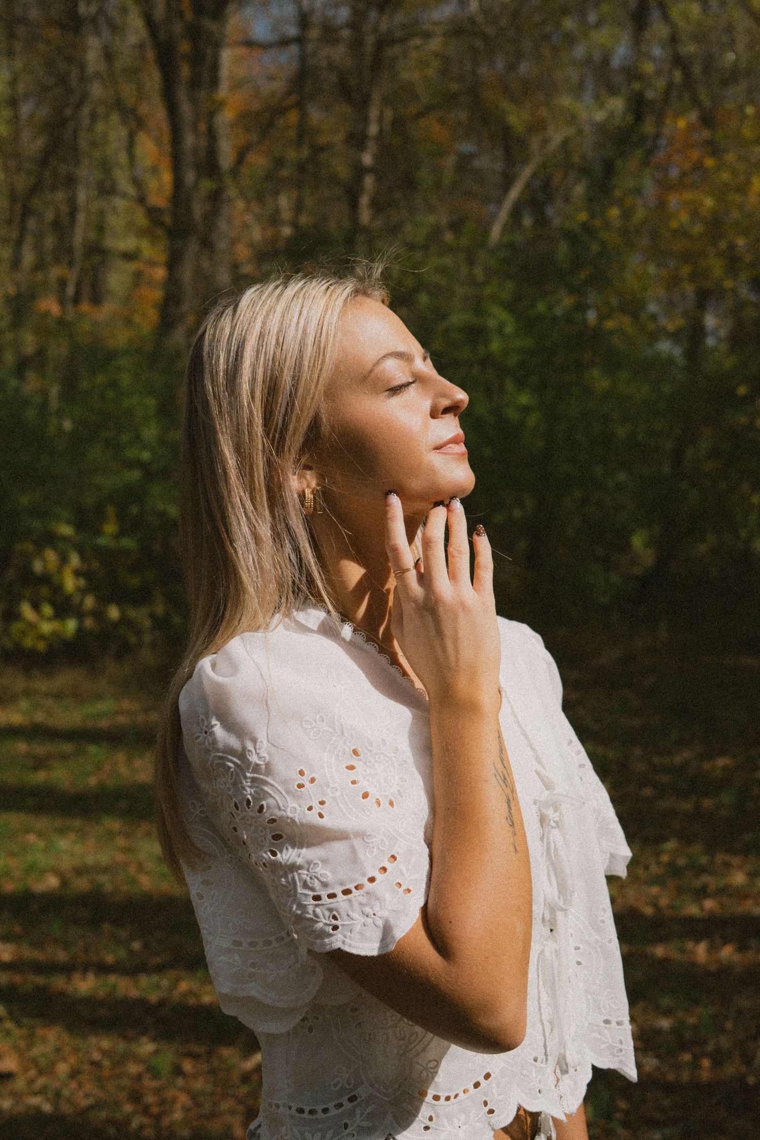 A person in a white eyelet lace dress poses thoughtfully against an autumn forest backdrop.