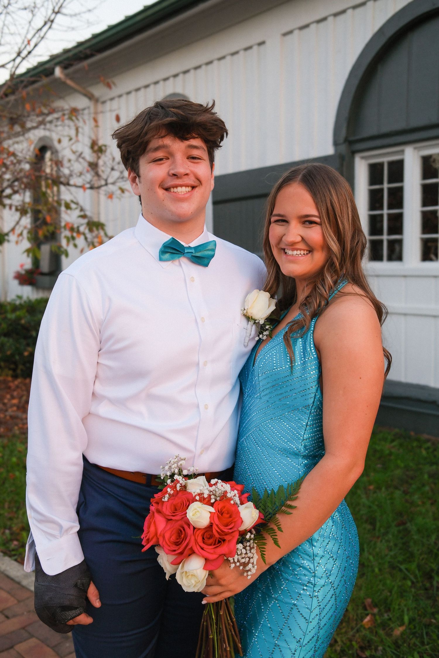 A couple in formal attire poses together at a special event, with the woman wearing a turquoise dress and holding a bouquet.