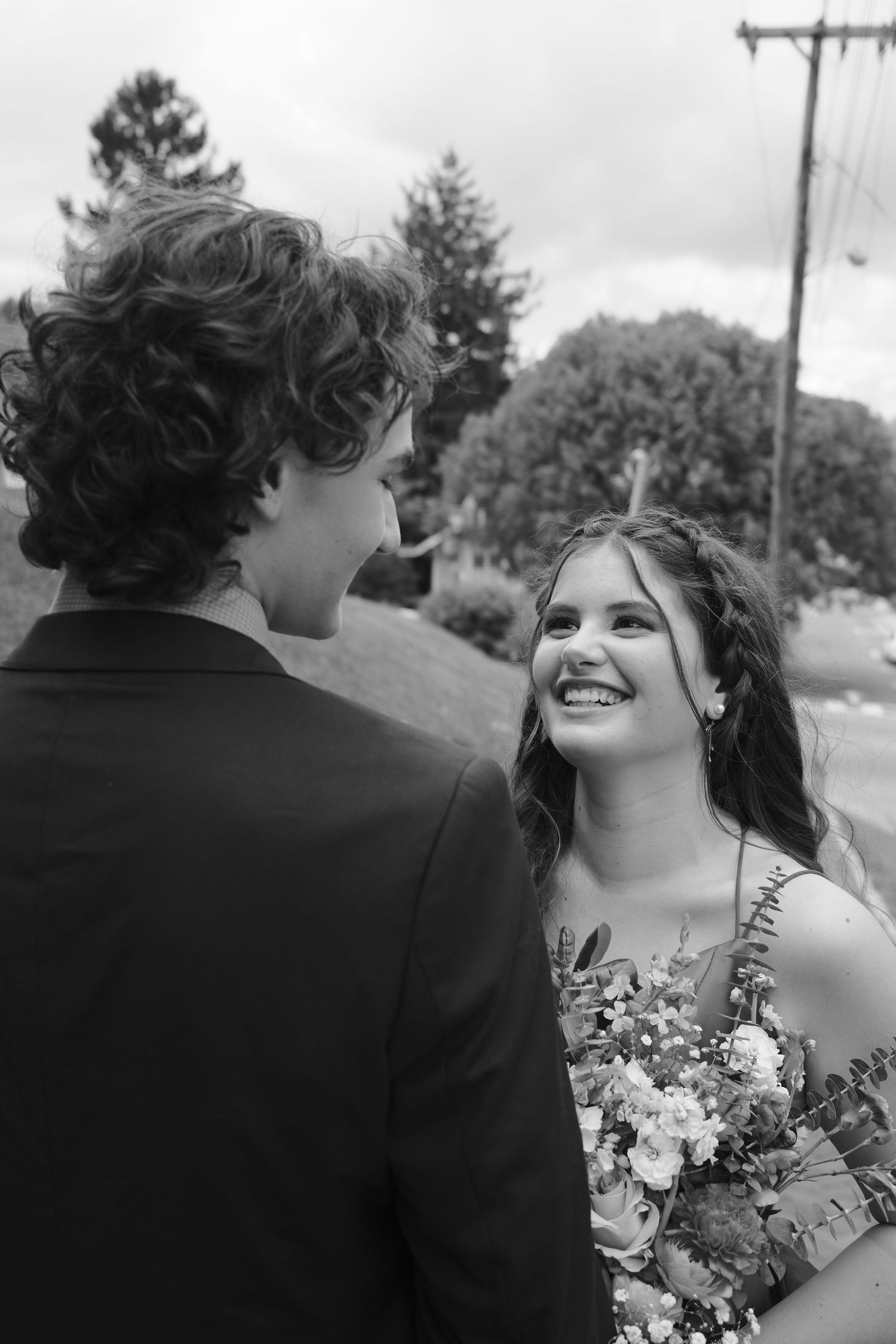 A joyful bride in a floral dress holds a white bouquet while looking up with a bright smile in a black and white photo.