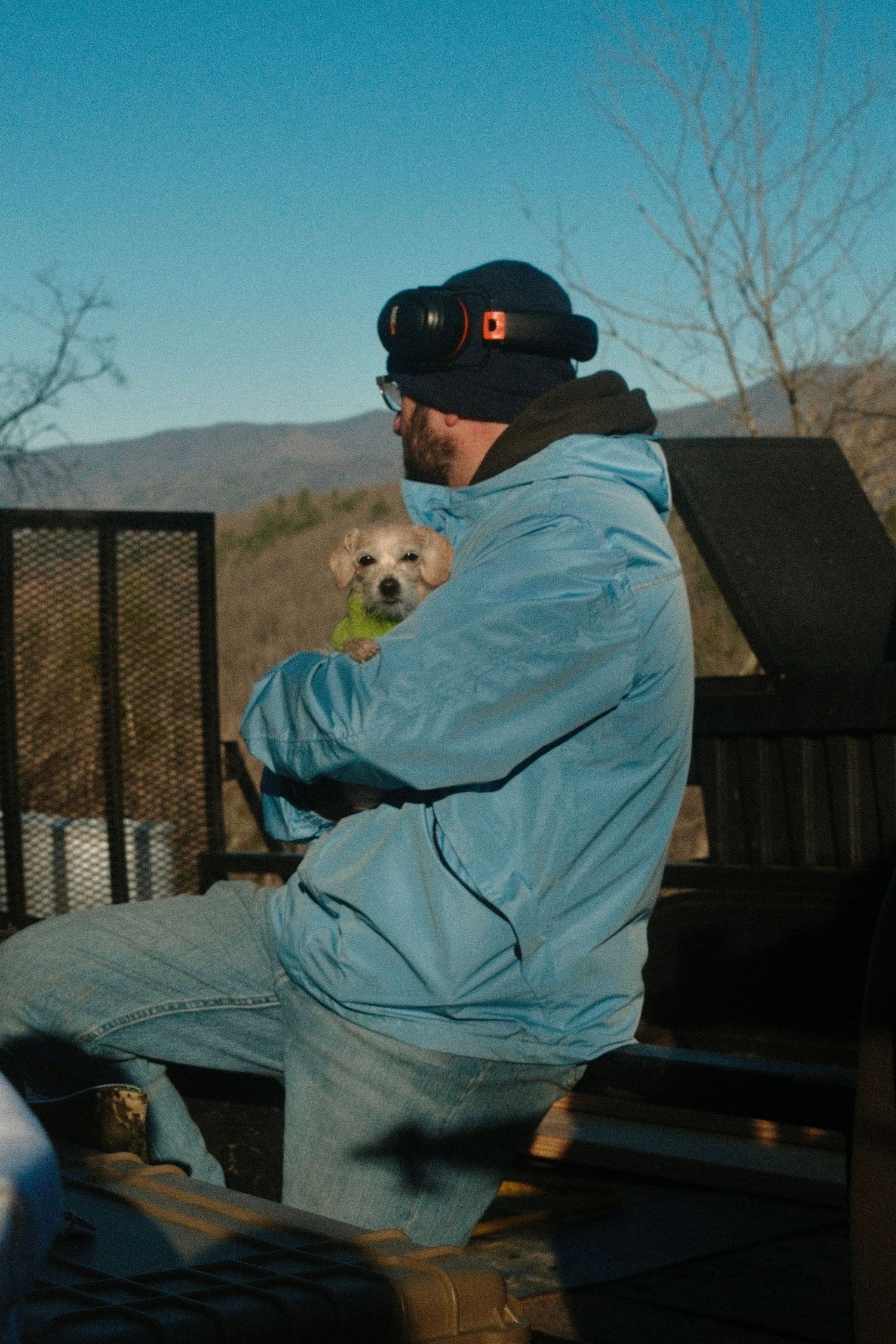 A person in a denim jacket sits on a bench outdoors during a sunny winter day with bare trees in the background.
