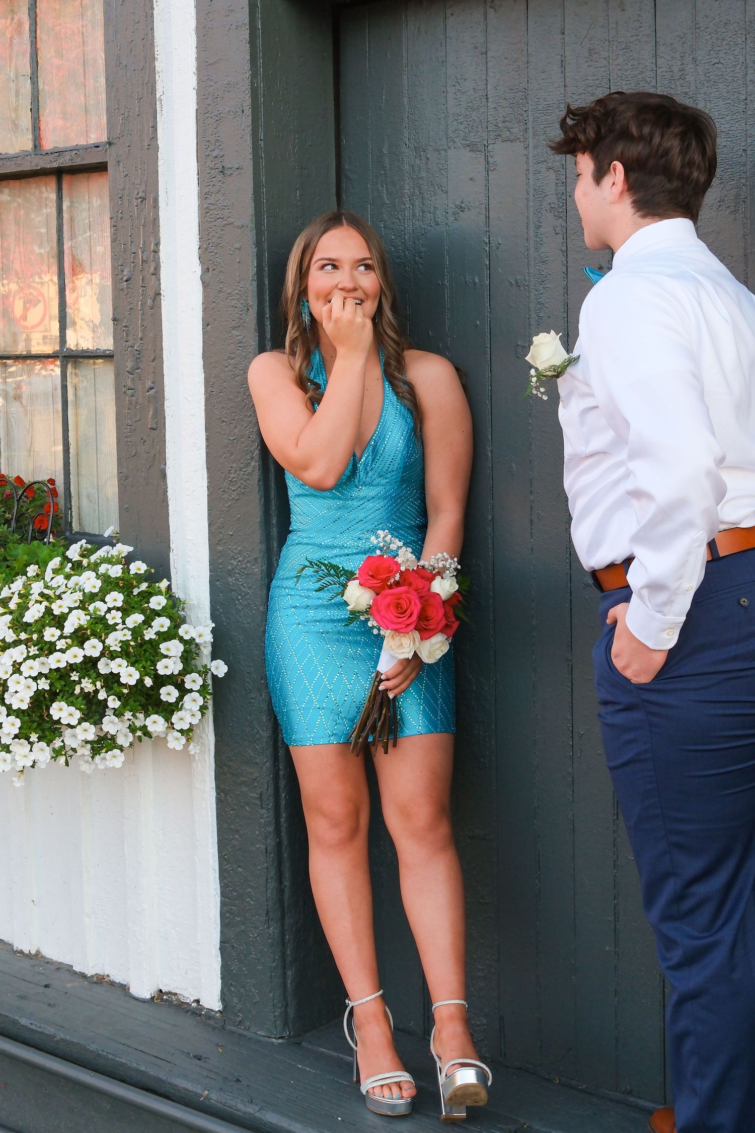 A couple in turquoise and navy outfits poses playfully against a dark wall with white flowers in the background.