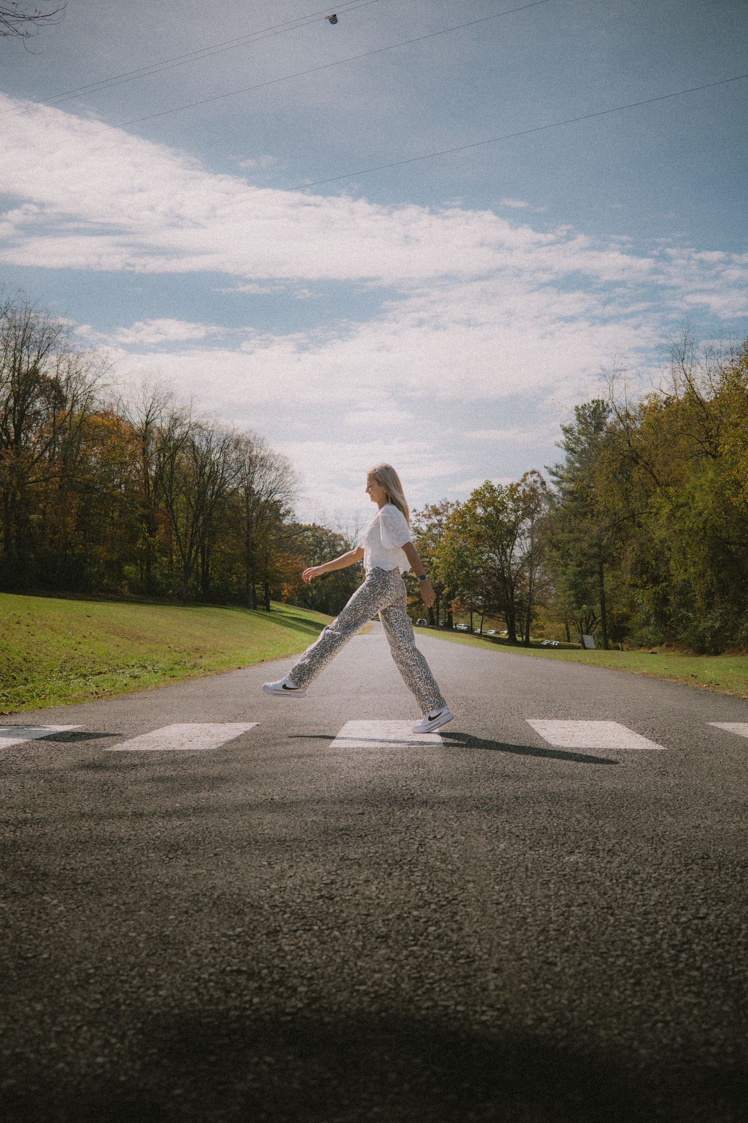 Person in white and leopard print performs dance moves on an empty road with trees in the background.
