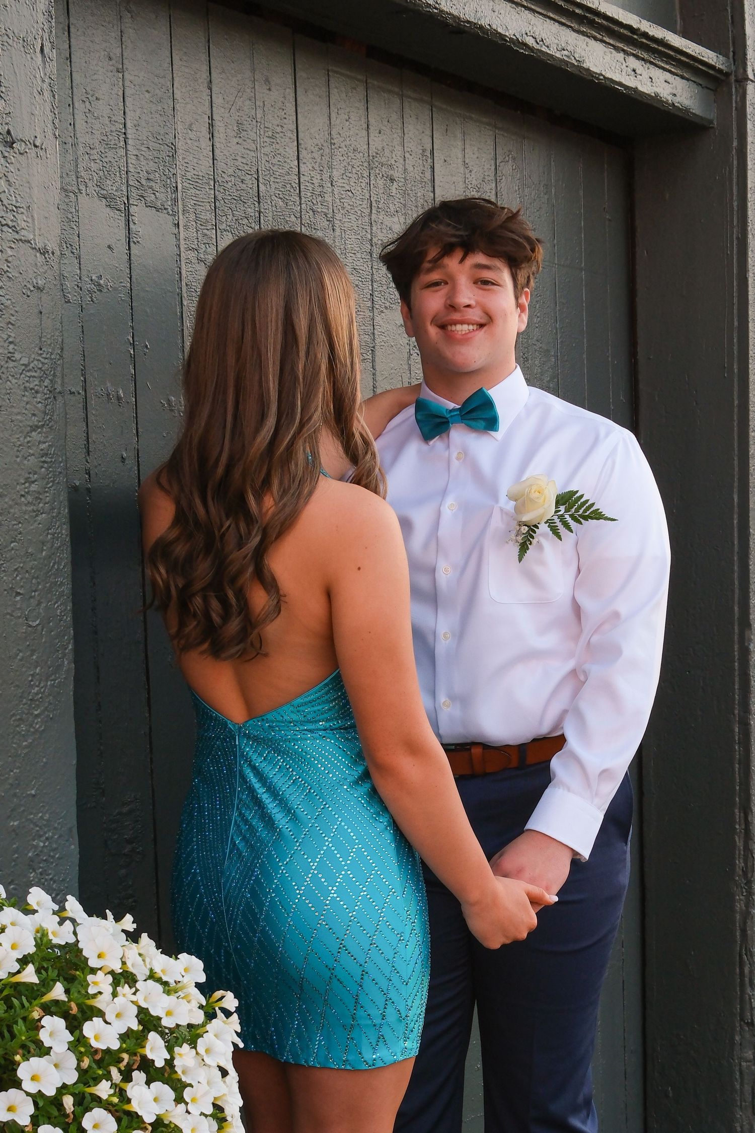 A couple in formal attire pose together against a gray wall with white flowers, the woman wearing a turquoise dress.