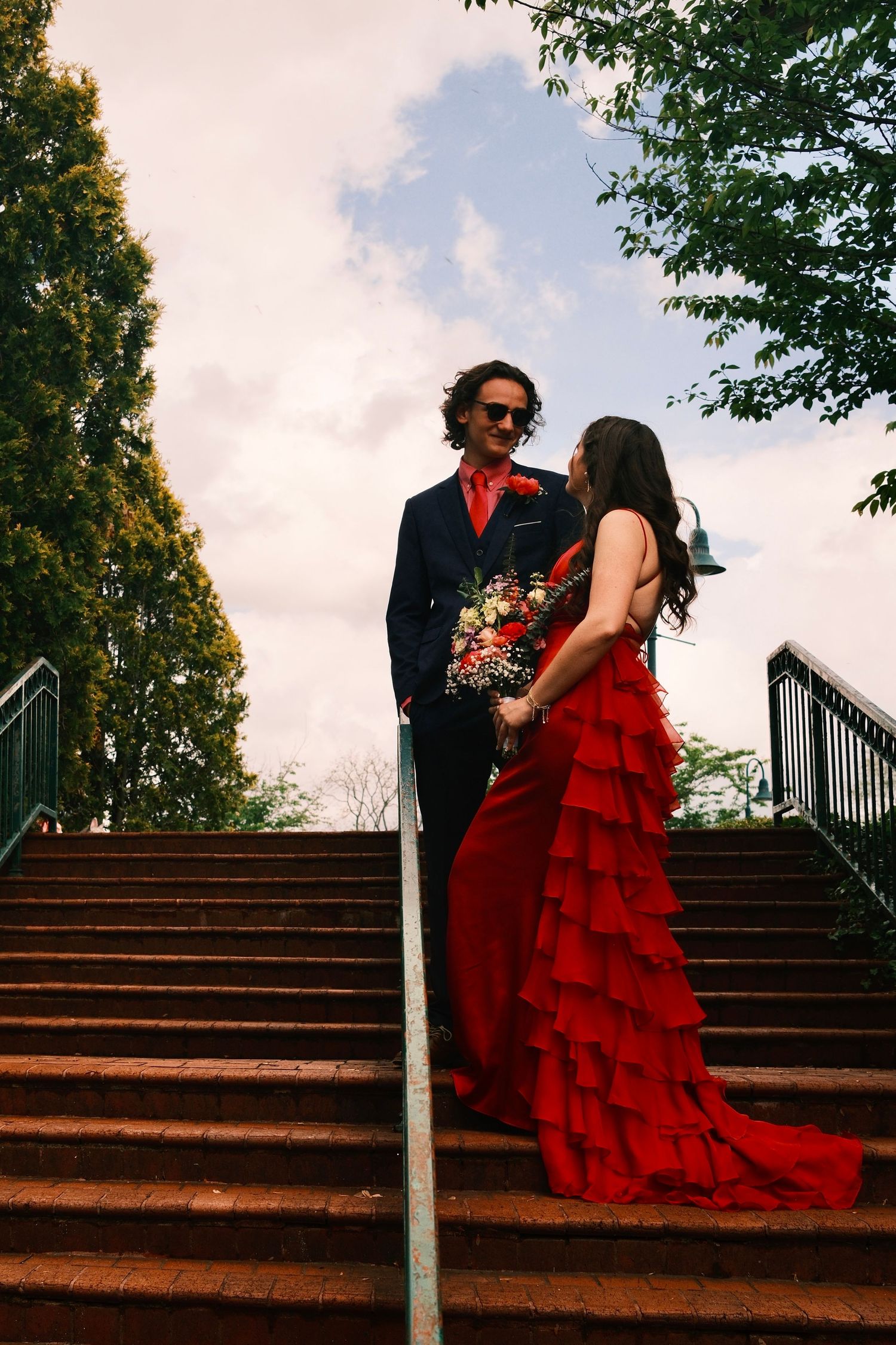 A couple in red and navy formal wear pose together on outdoor brick steps.