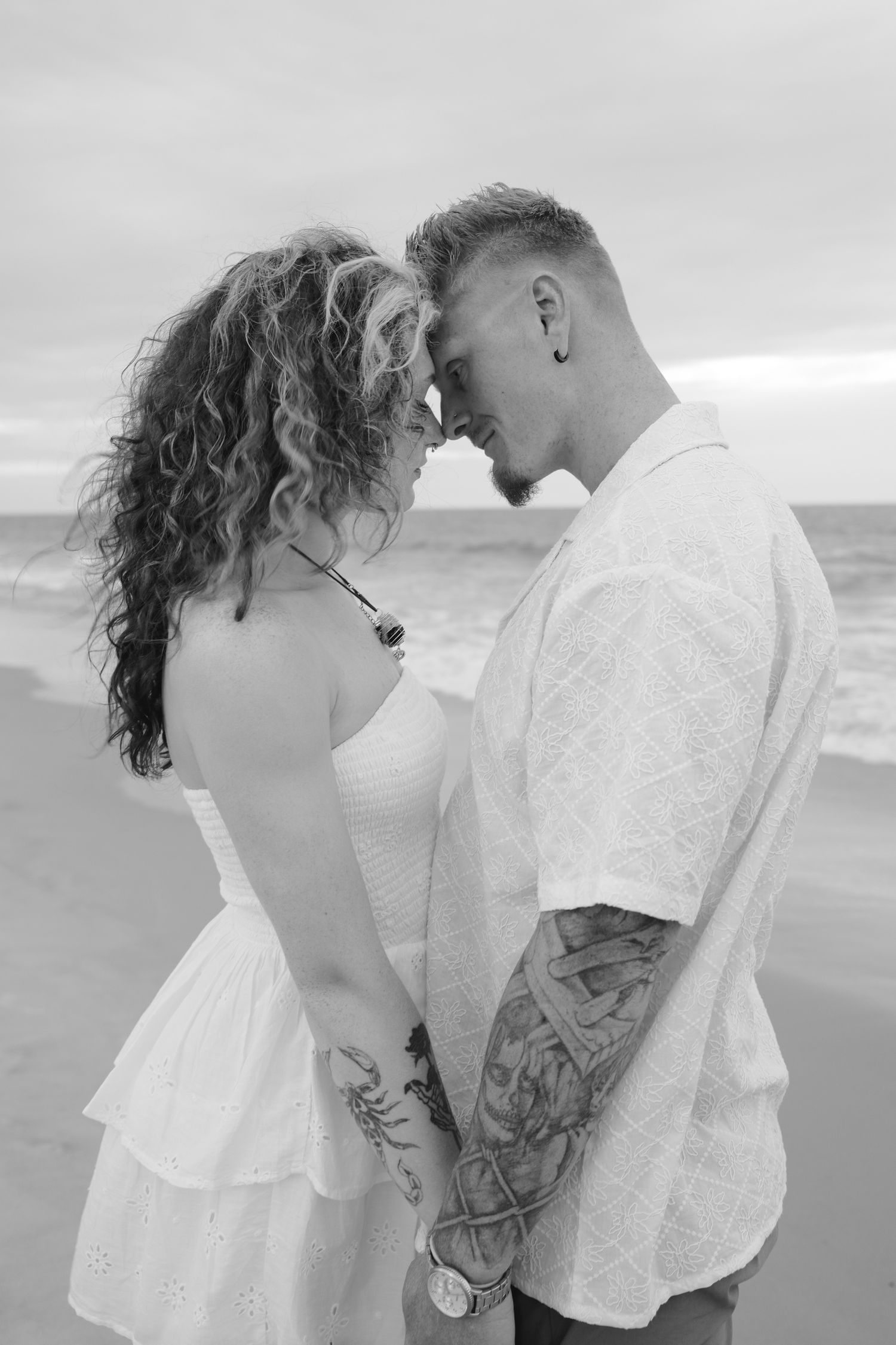 A romantic black and white beach photoshoot series showing tender moments between a couple in white outfits by the ocean.