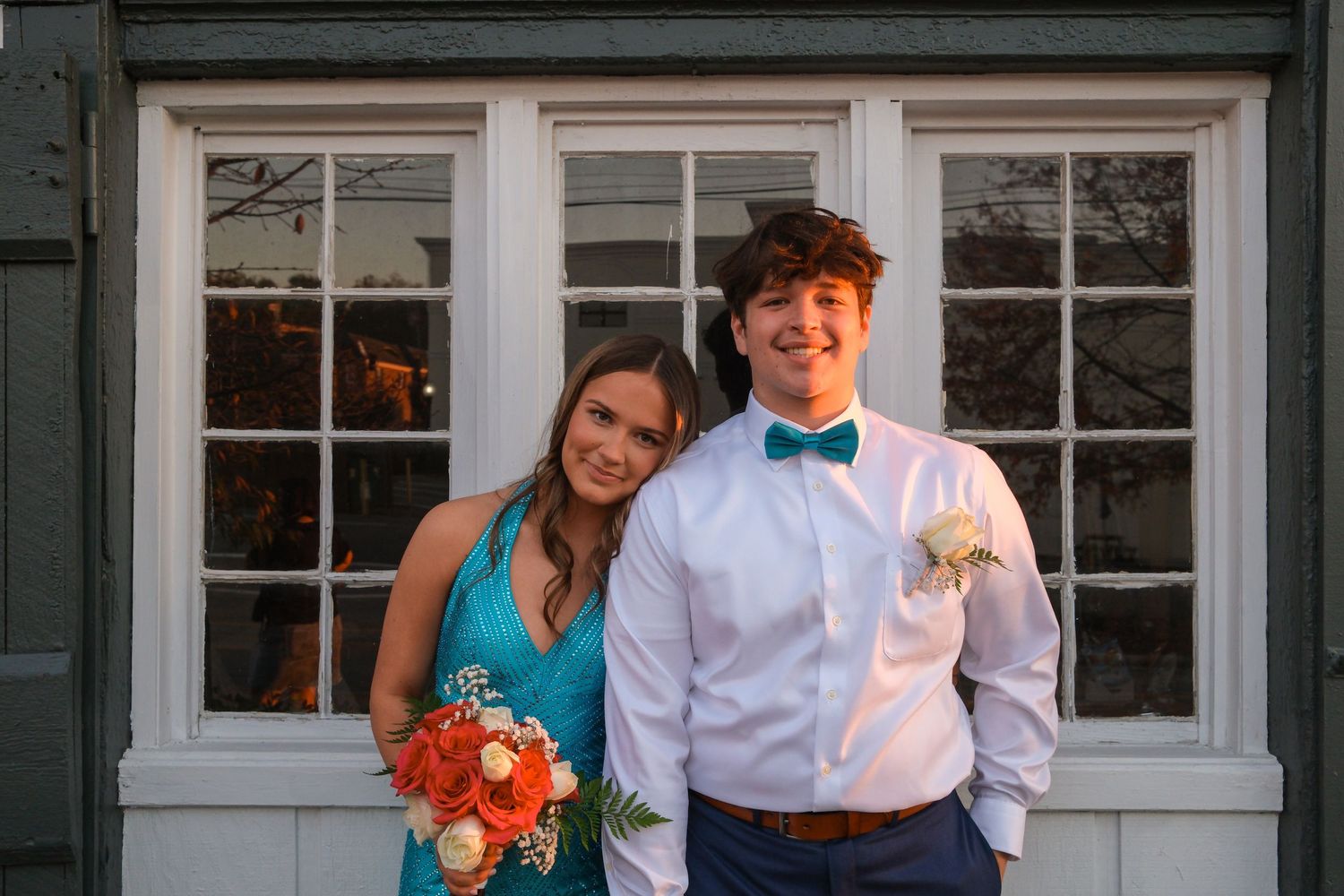 Couple in formal wear smiles together in front of white building with windows.