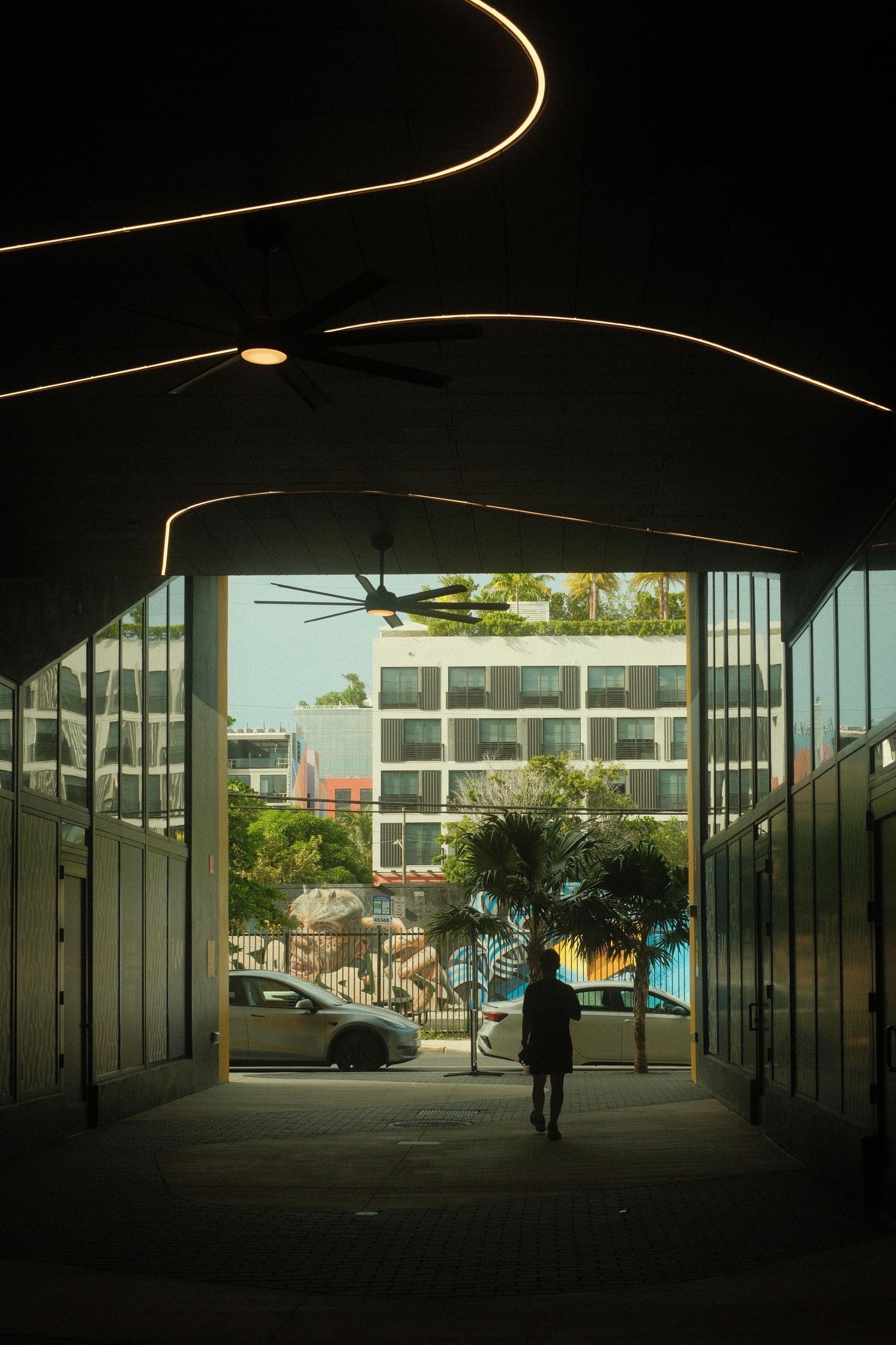 A silhouetted figure walks through a dark covered walkway with curved architectural lines and natural light at the end.