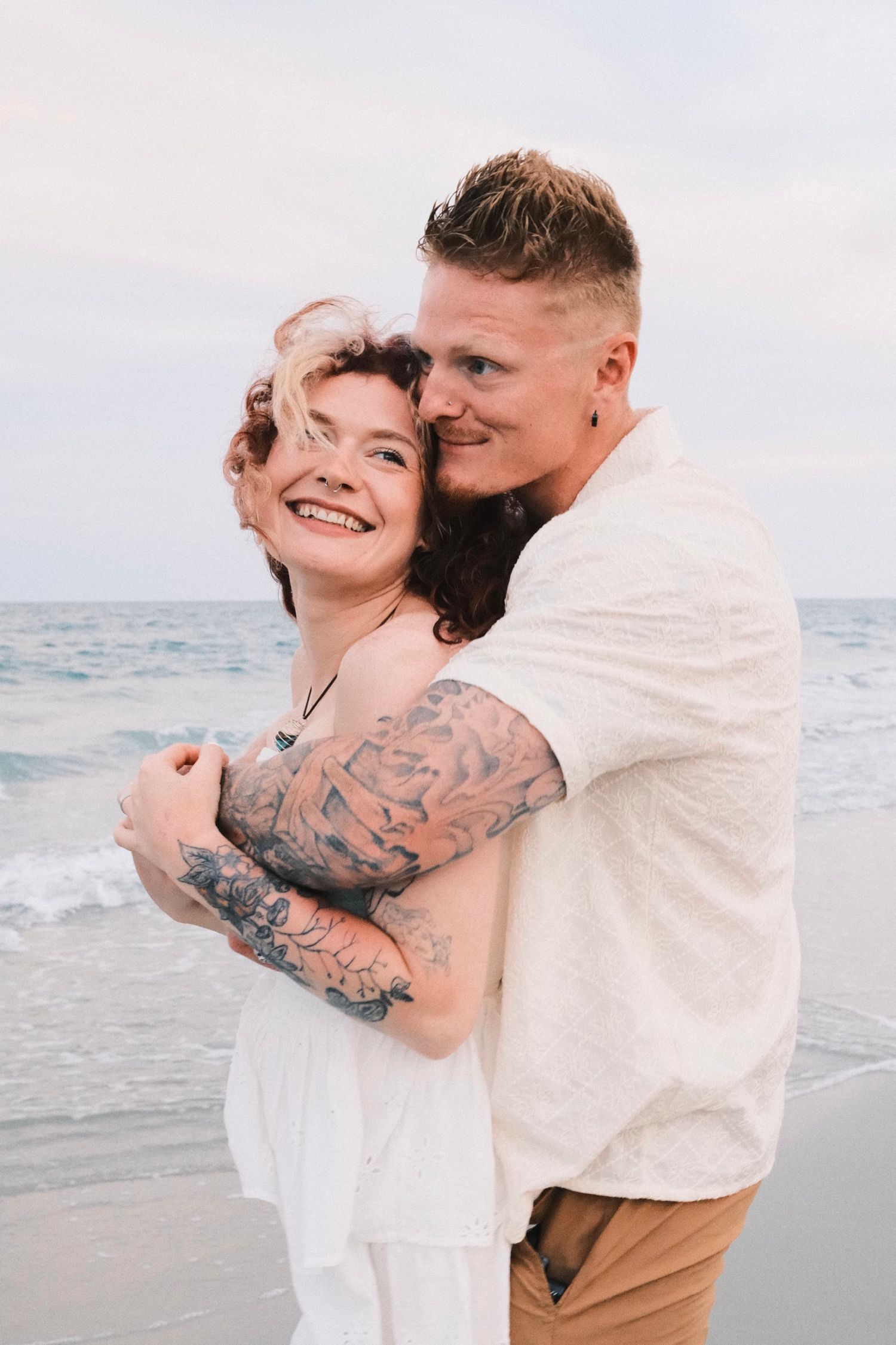 A romantic couple embraces and laughs together during a beach photoshoot at sunset.