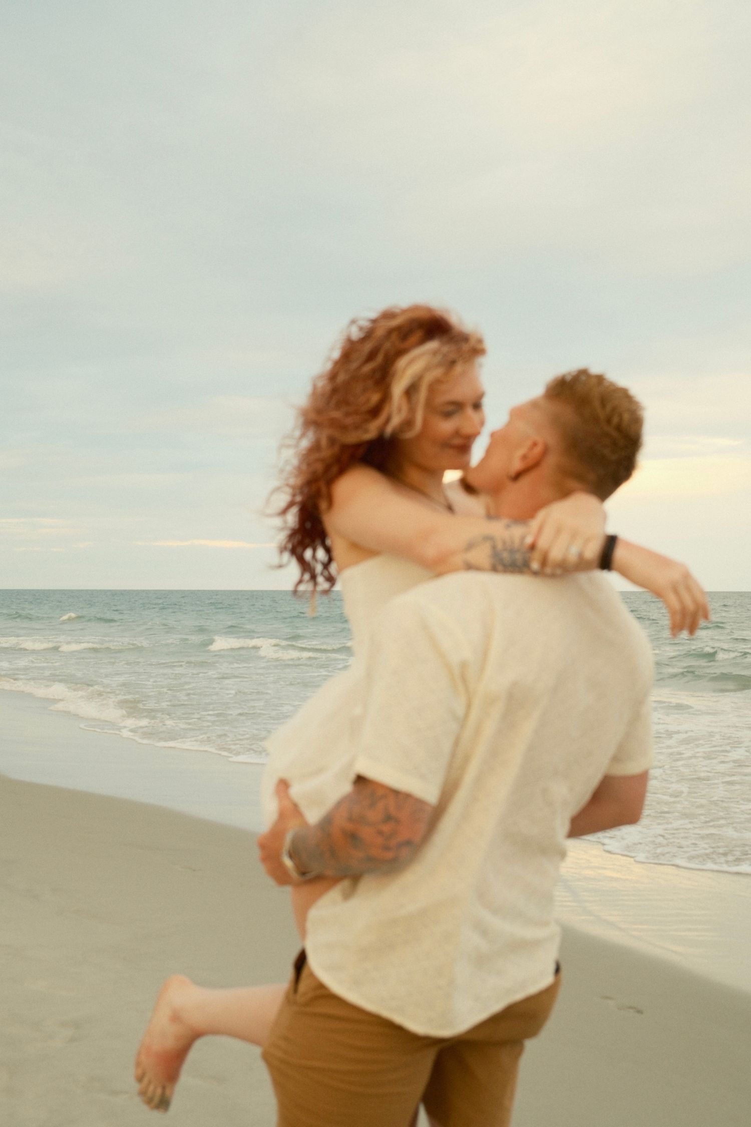 Romantic couple embracing and kissing on a sandy beach at sunset with ocean waves in the background.