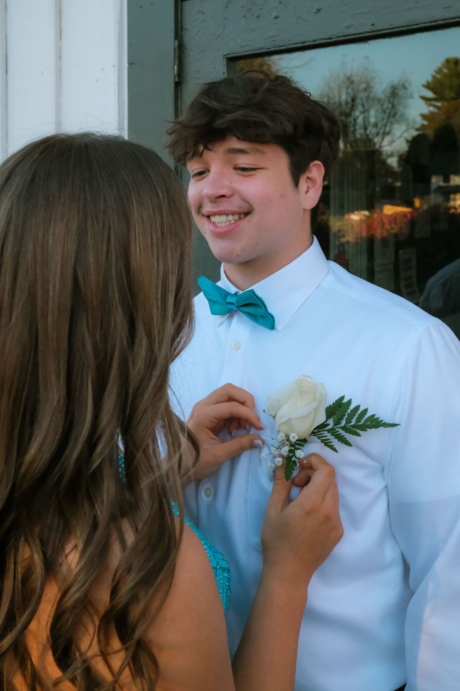 A couple in formal attire prepare for a dance, with someone pinning a white boutonniere to a turquoise bowtie.