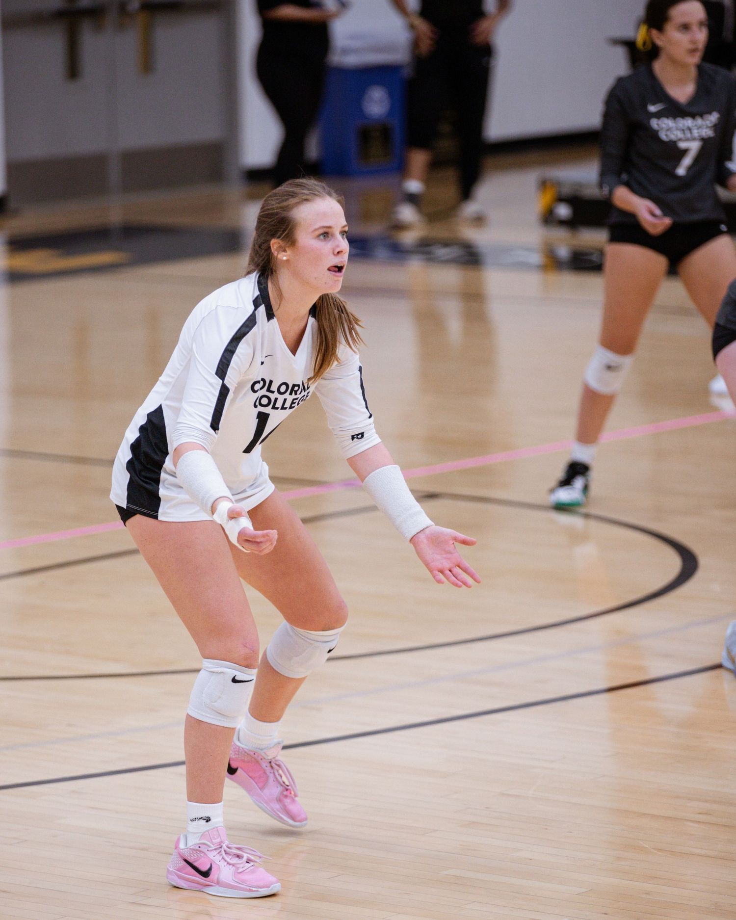 Volleyball player in white uniform crouches low in defensive position on indoor court during game play.