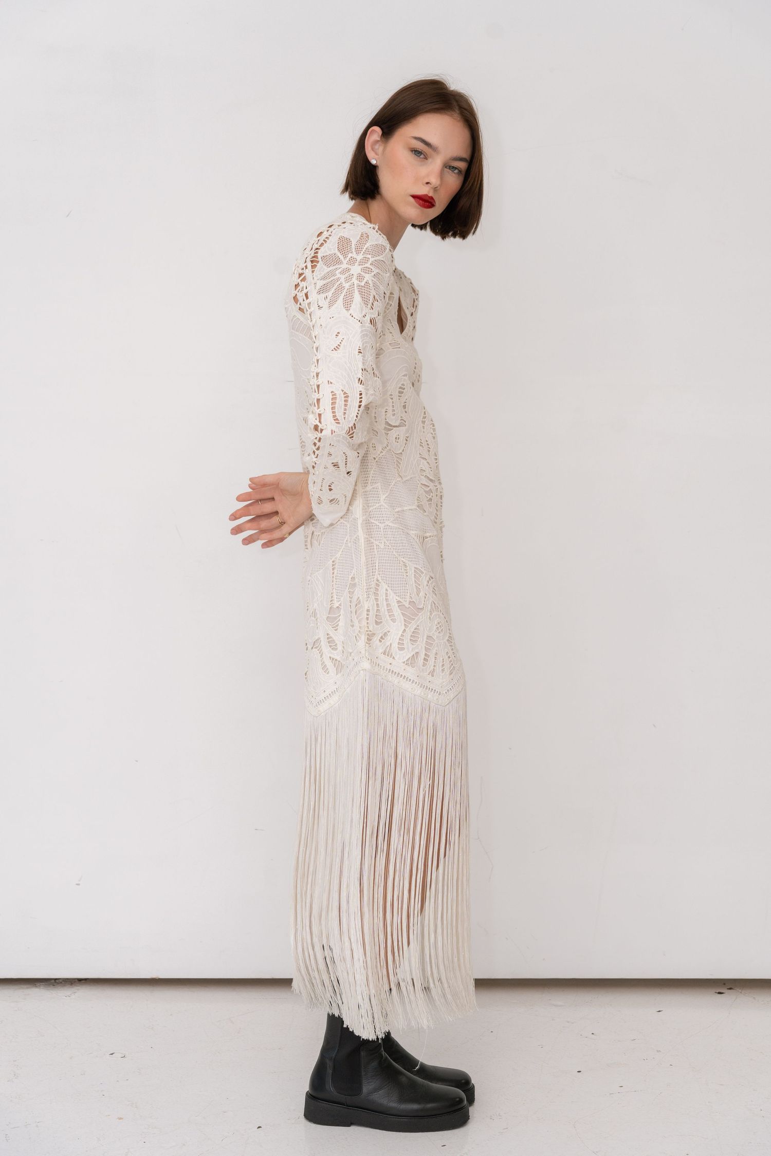 Female model standing in white studio wearing flowing white dress, arms behind back, fashion editorial by Sadie Sanders