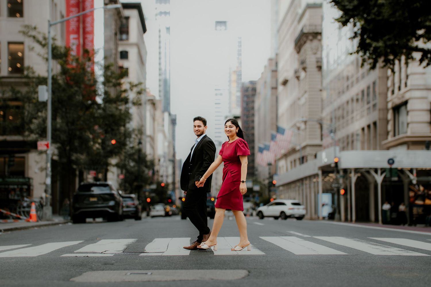 Couple walking across urban crosswalk with red accents in their outfits and architecture visible in background.