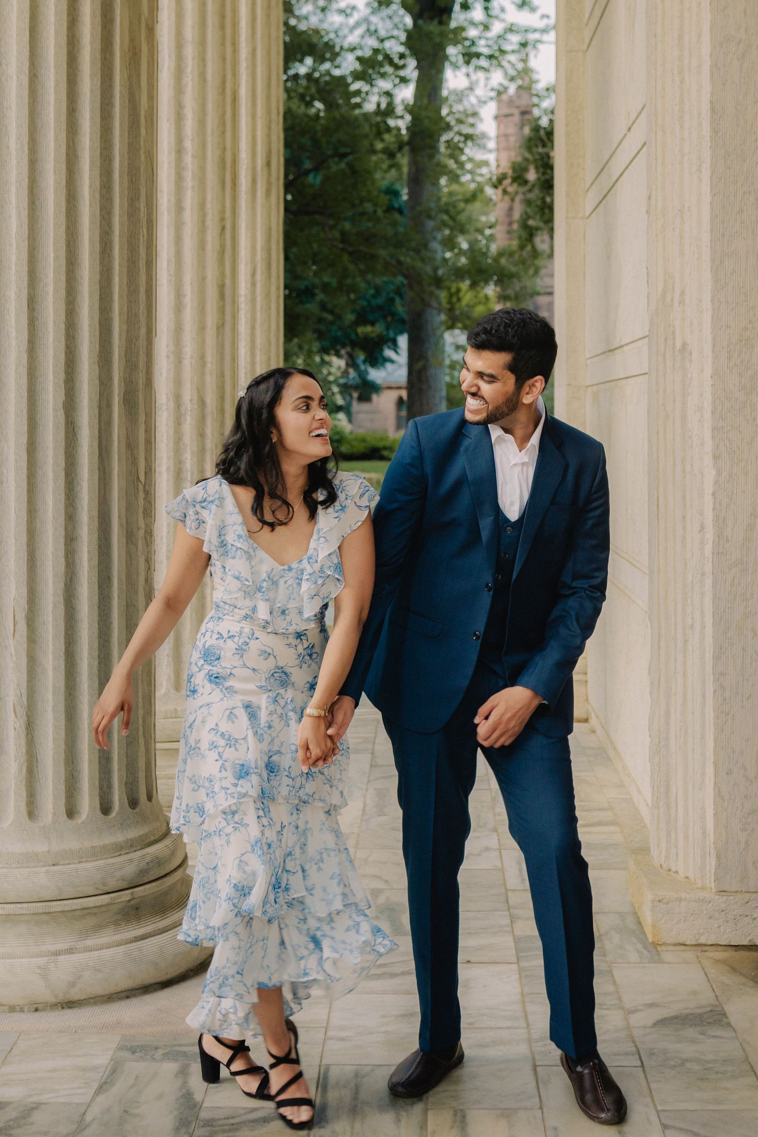 A couple in blue dress and navy suit playfully poses between classical stone columns in an engagement photoshoot.