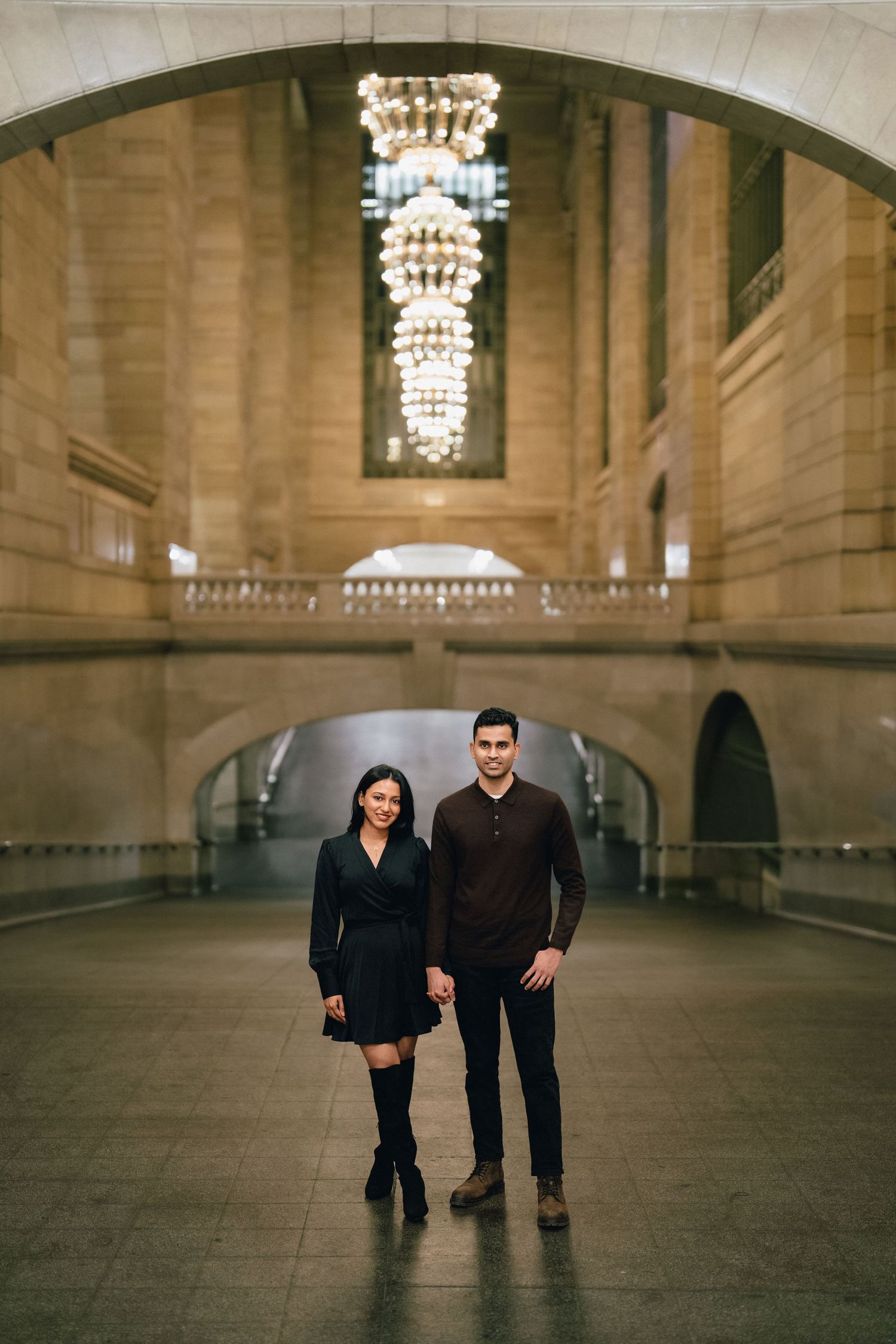 Two people walk together through the grand interior of Grand Central Terminal with ornate chandeliers and stone archways.