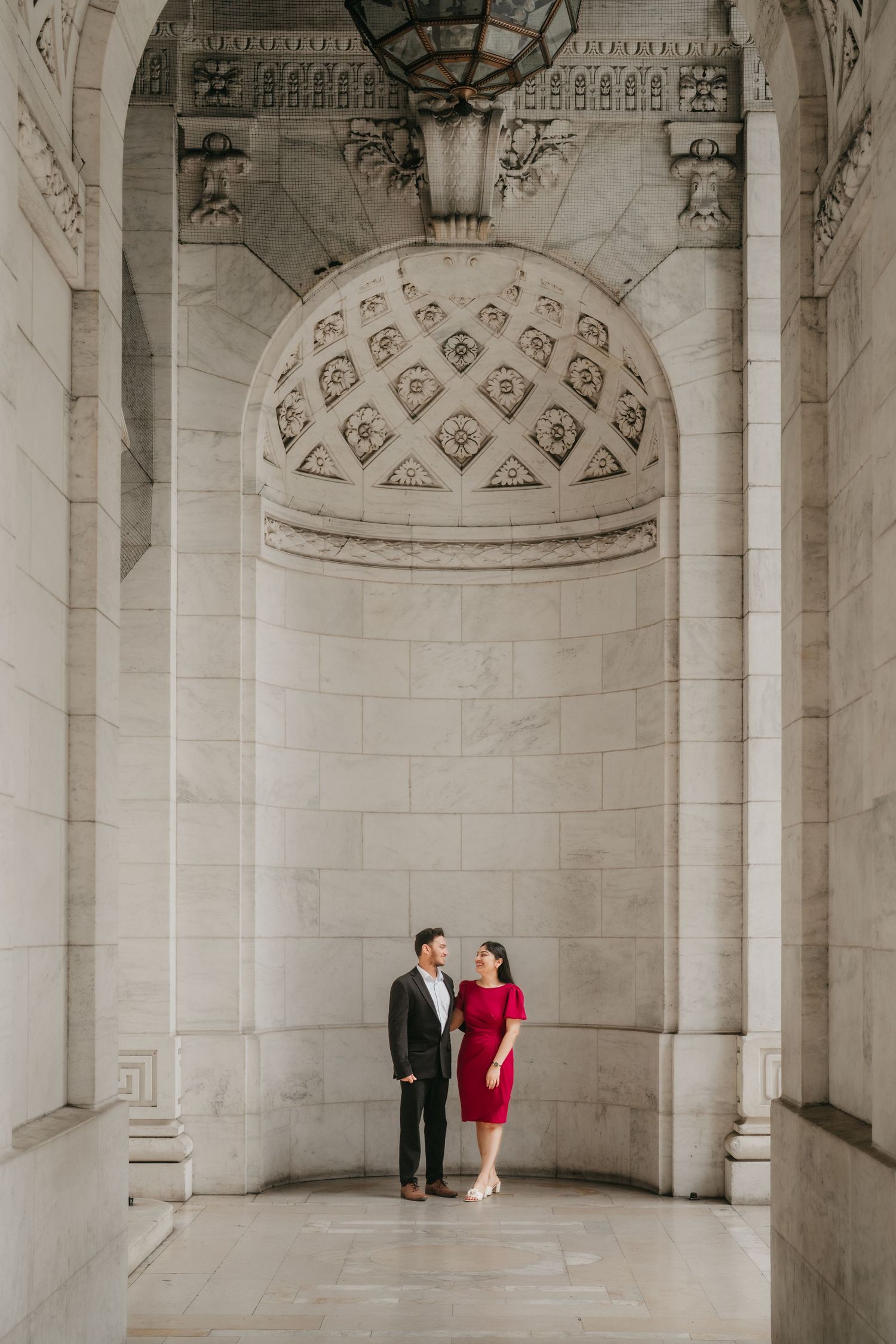 A couple in red attire shares a romantic moment in an elegant white marble colonnade with ornate architectural details.
