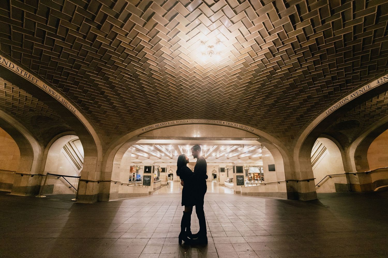 A romantic couple embraces in an arched brick tunnel with warm lighting creating a dramatic silhouette effect.