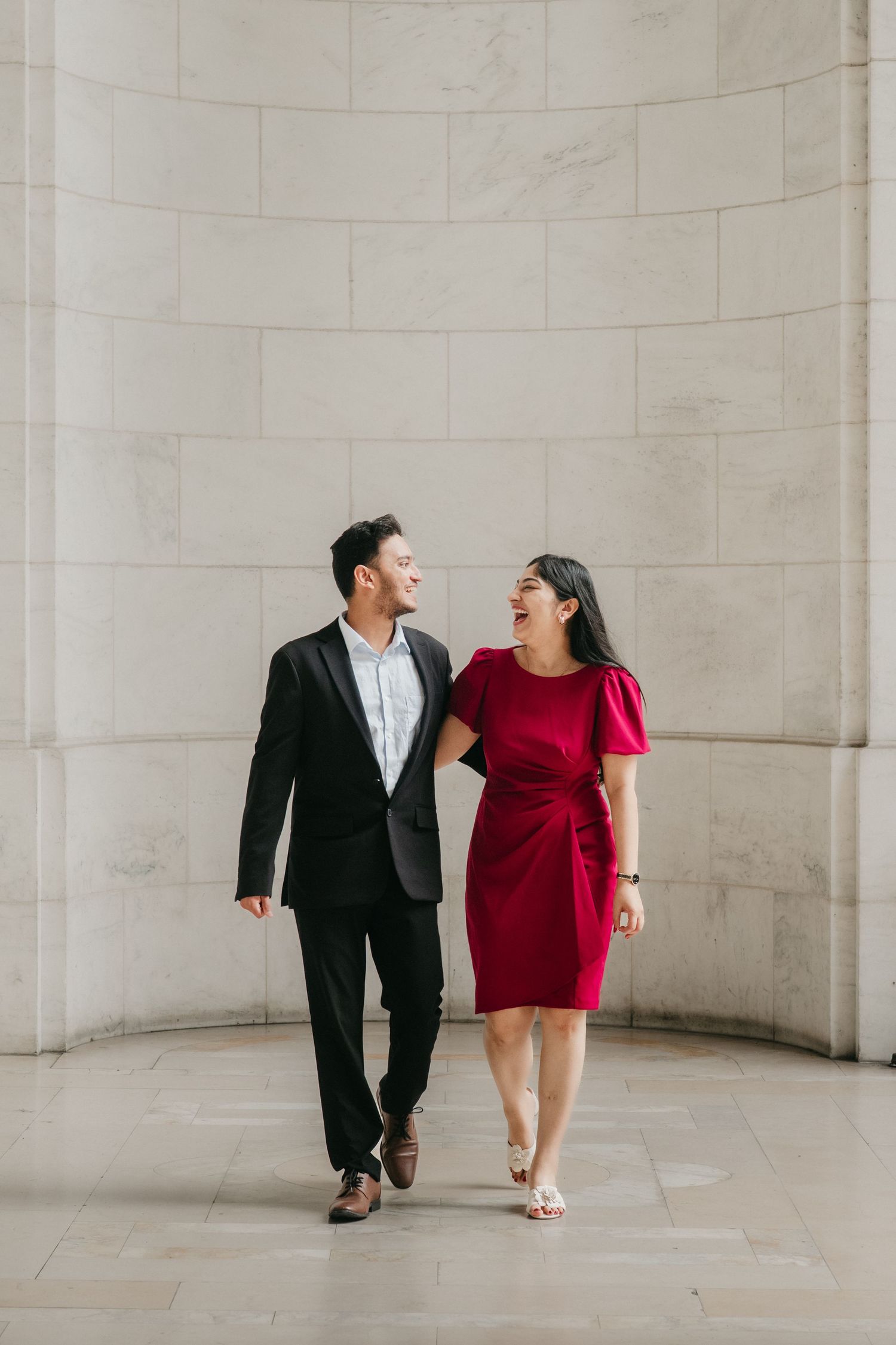 A couple in formal attire shares playful moments in a minimalist hallway with white stone walls.