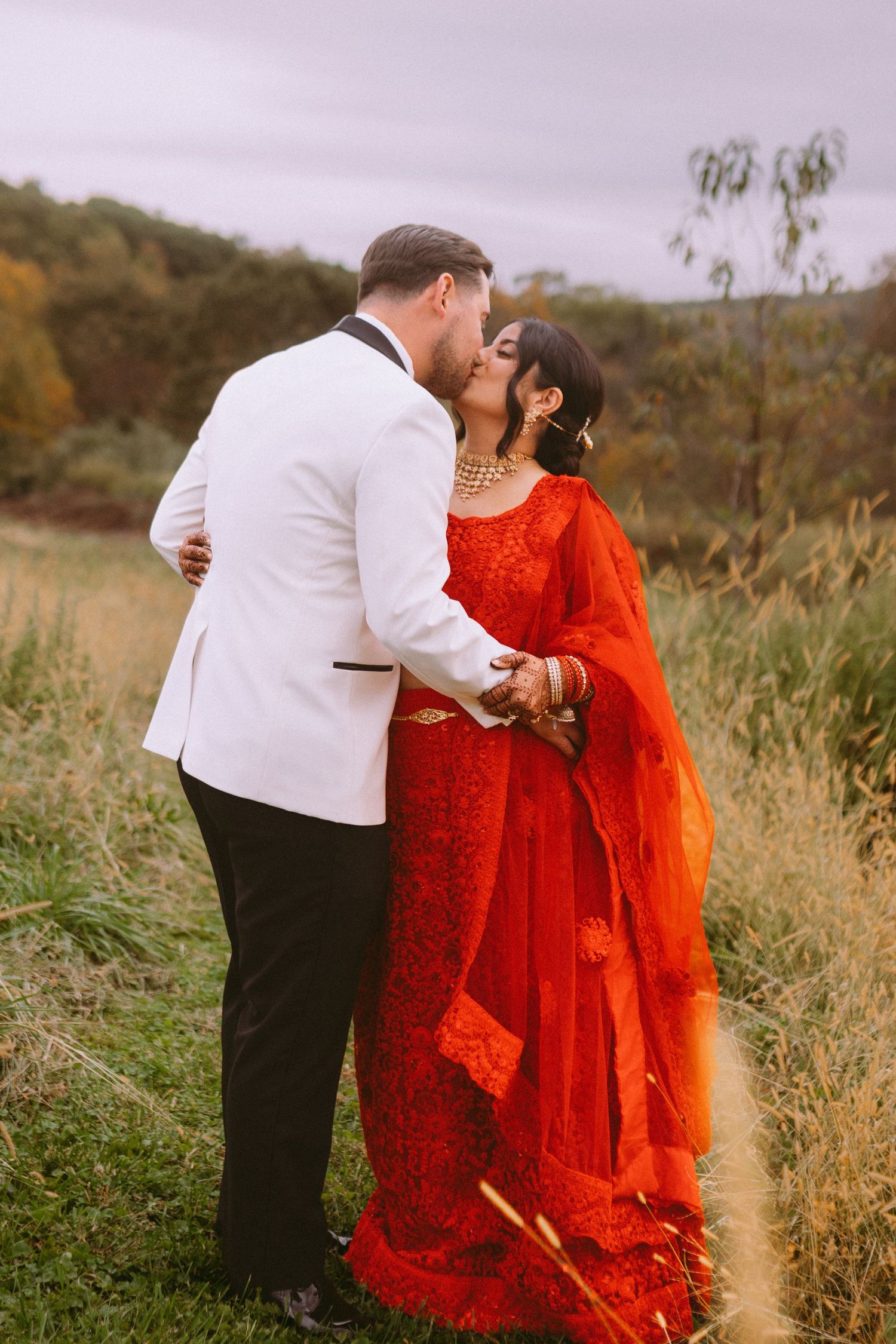 Romantic couple embracing in a field wearing red and white formal attire at sunset.
