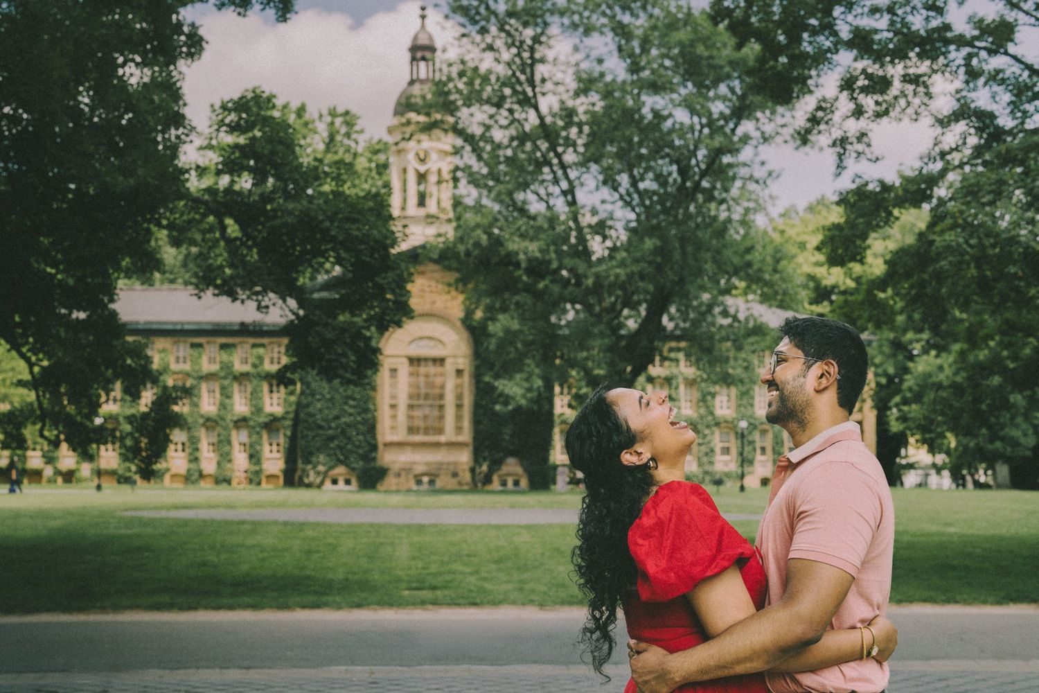 A couple embraces in a romantic moment while walking through a scenic park with historic buildings in the background.