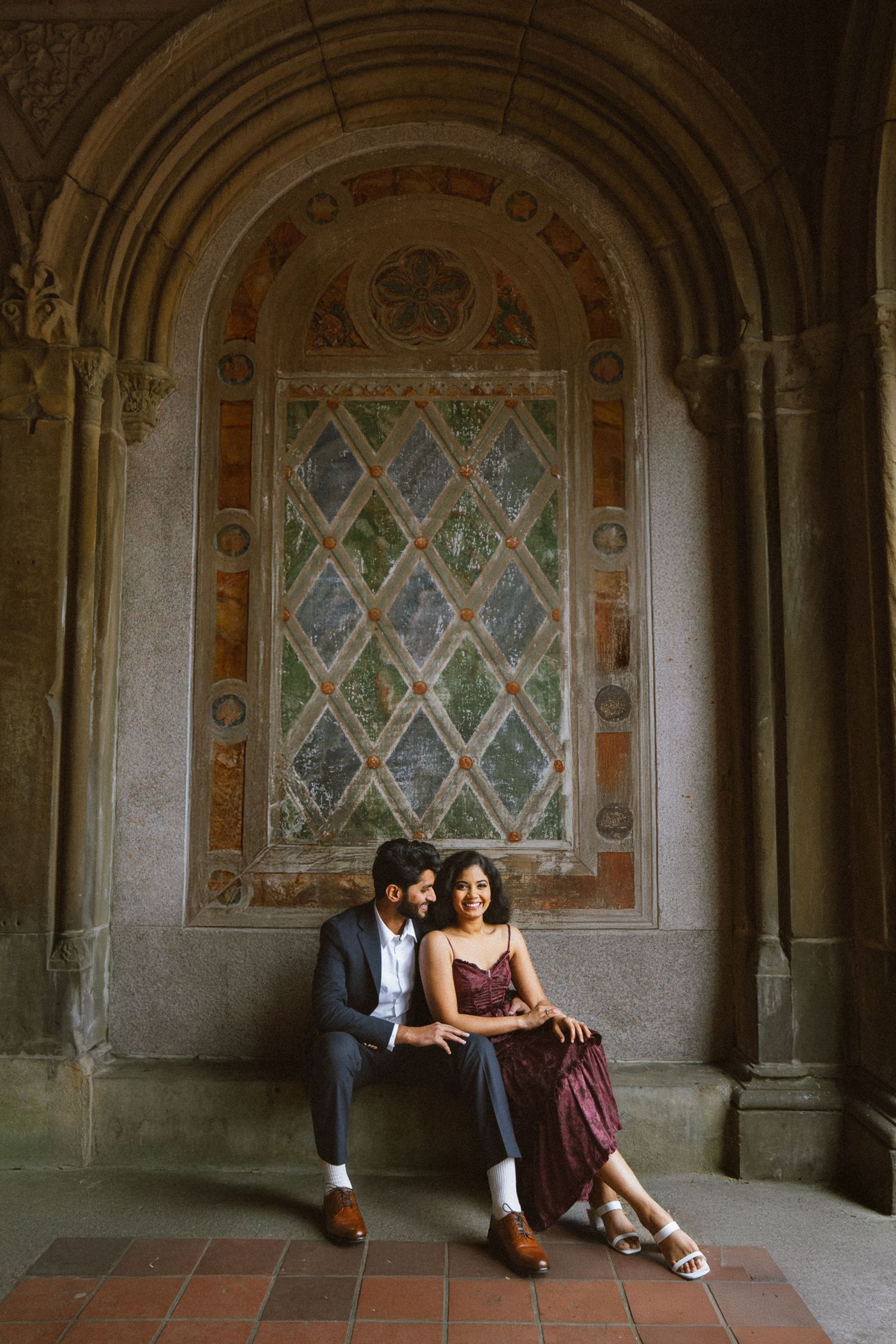 Couple sitting in historic stone archway with stained glass windows and decorative details.