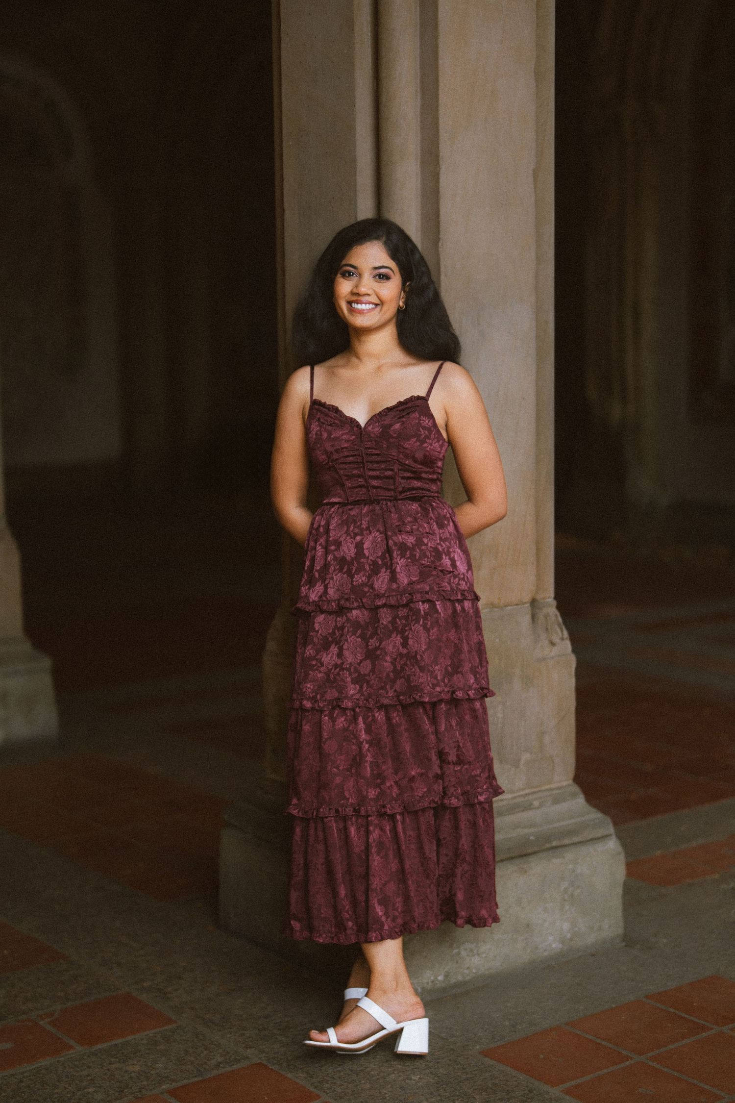 Woman in burgundy tiered lace dress and white sandals poses elegantly by stone column in outdoor setting.