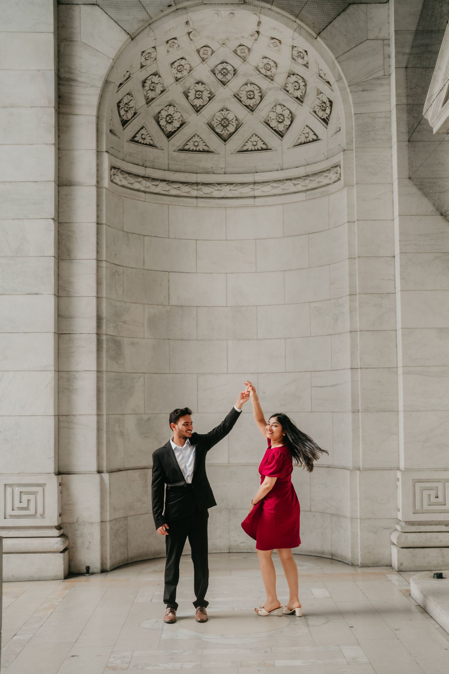 Couple dances playfully under ornate domed archways in a grand white stone building interior.