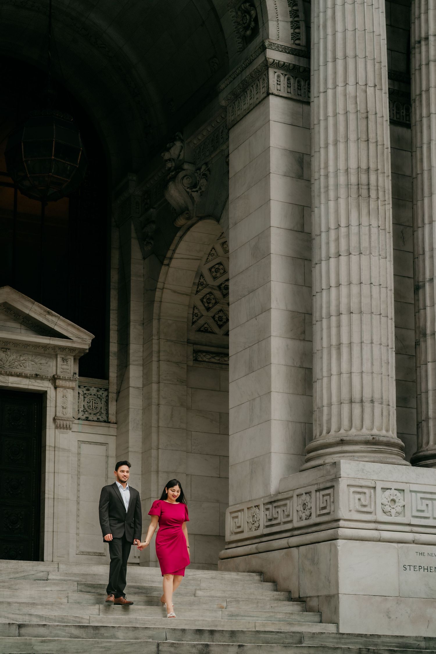 A couple stands on the grand stone steps of a neoclassical building with ornate columns and architectural details.