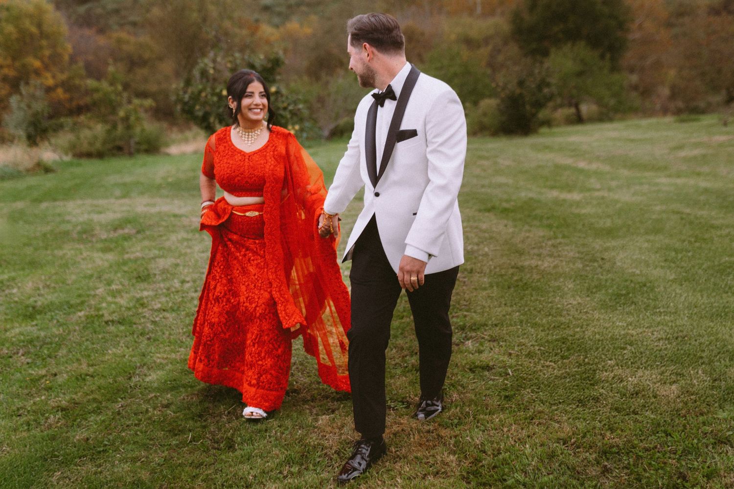 A couple walks hand in hand through a grassy field, the person in a bright red dress creating a striking contrast.