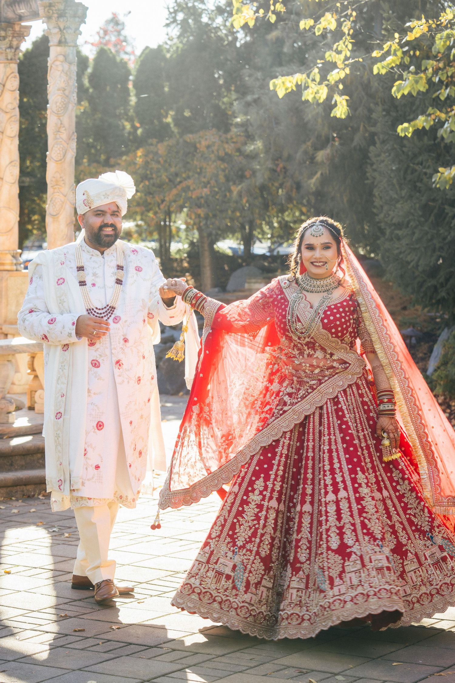 A couple in traditional Indian wedding attire twirling in sunlit outdoor setting.