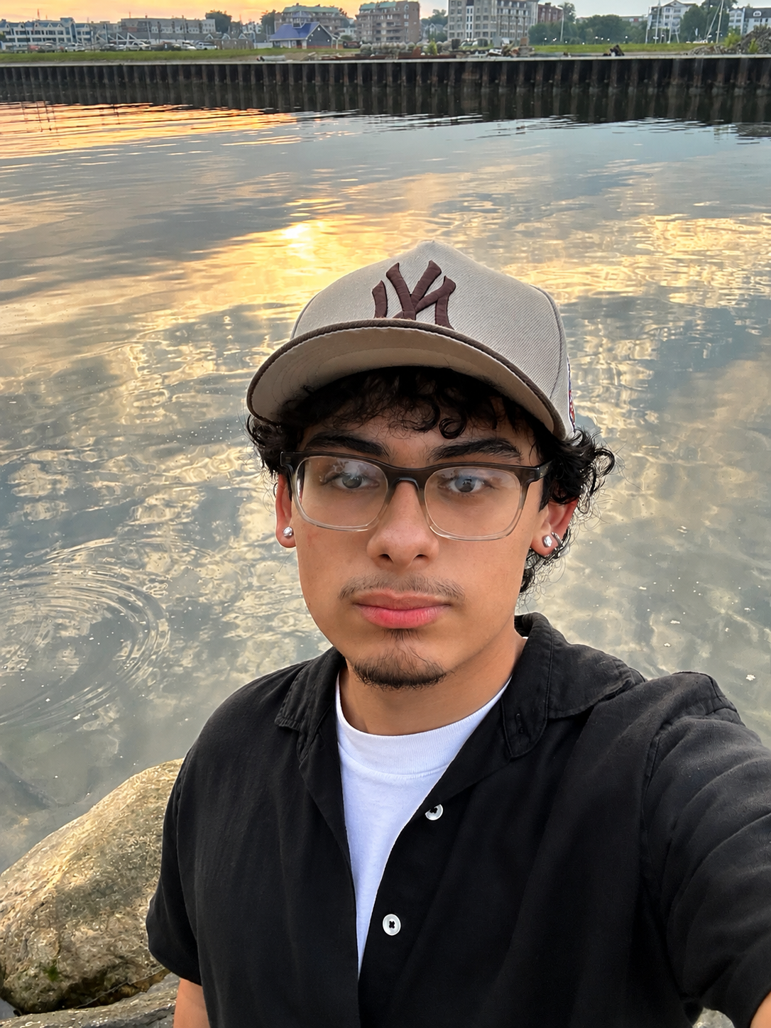 Young man wearing NY Yankees cap and glasses takes a selfie by a reflective waterway at sunset.