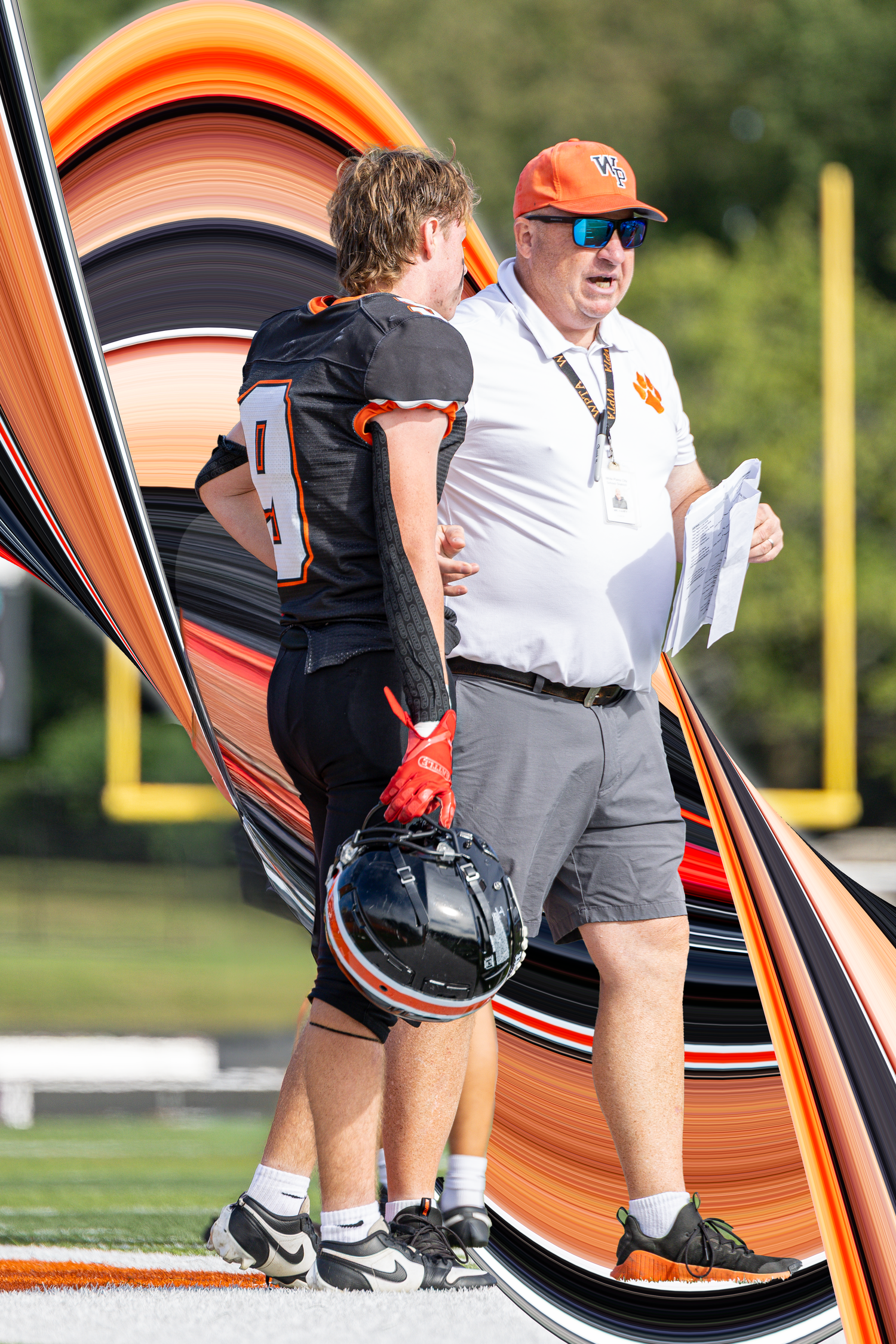 Football coach in orange cap reviews plays with a player in black uniform on a sunny sideline.
